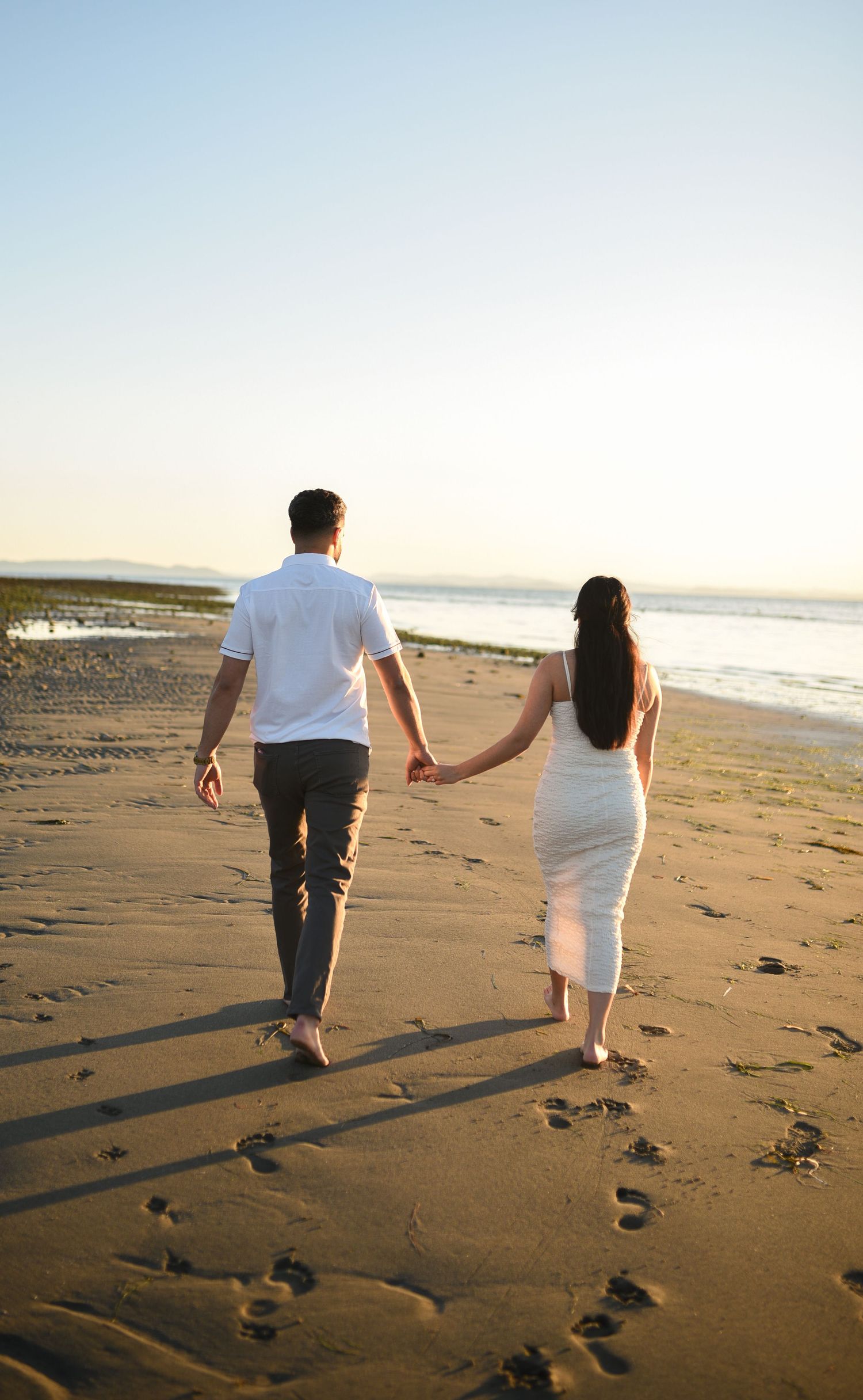 Couple holding hands walking barefoot on sandy beach at sunset, leaving footprints, woman in white dress, man in white shirt.