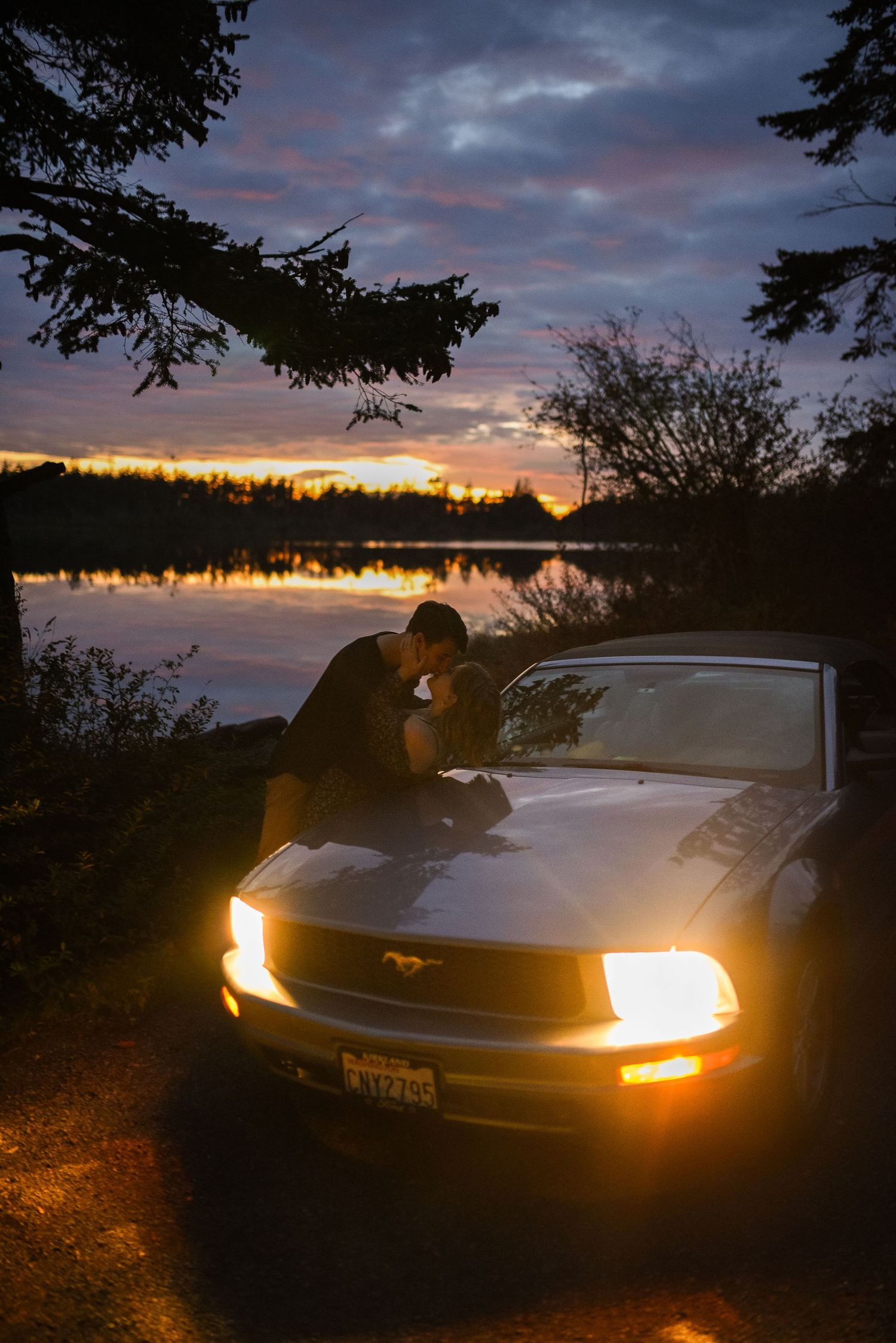 Couple kissing over the hood of a lit Ford Mustang at dusk with a colorful lake sunset reflection.