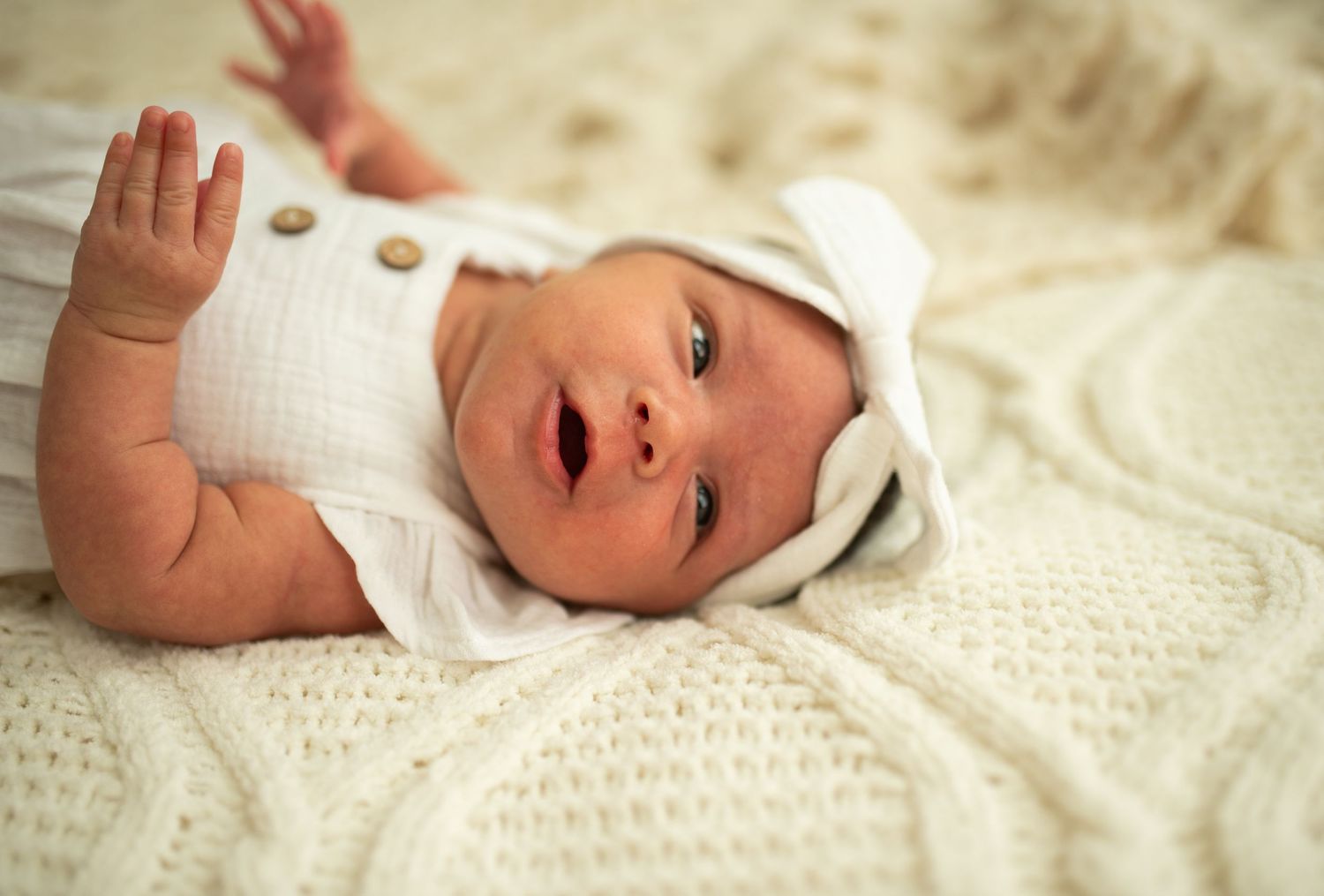 Alert newborn baby in white outfit and hat lying on a cream knit blanket looking up.