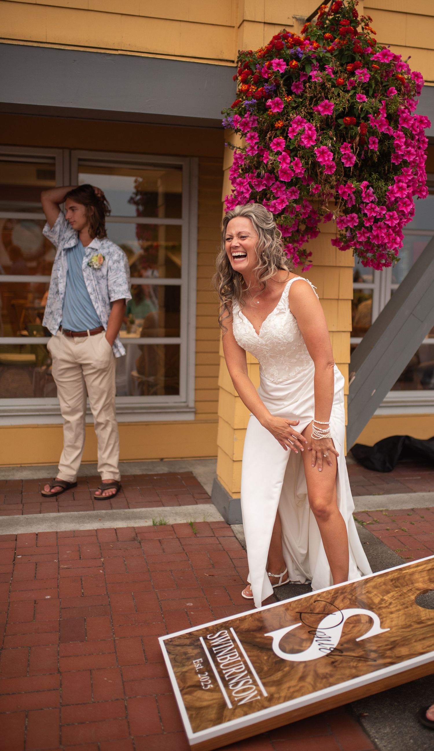Laughing bride in white gown plays cornhole outside a yellow building with pink hanging flowers.