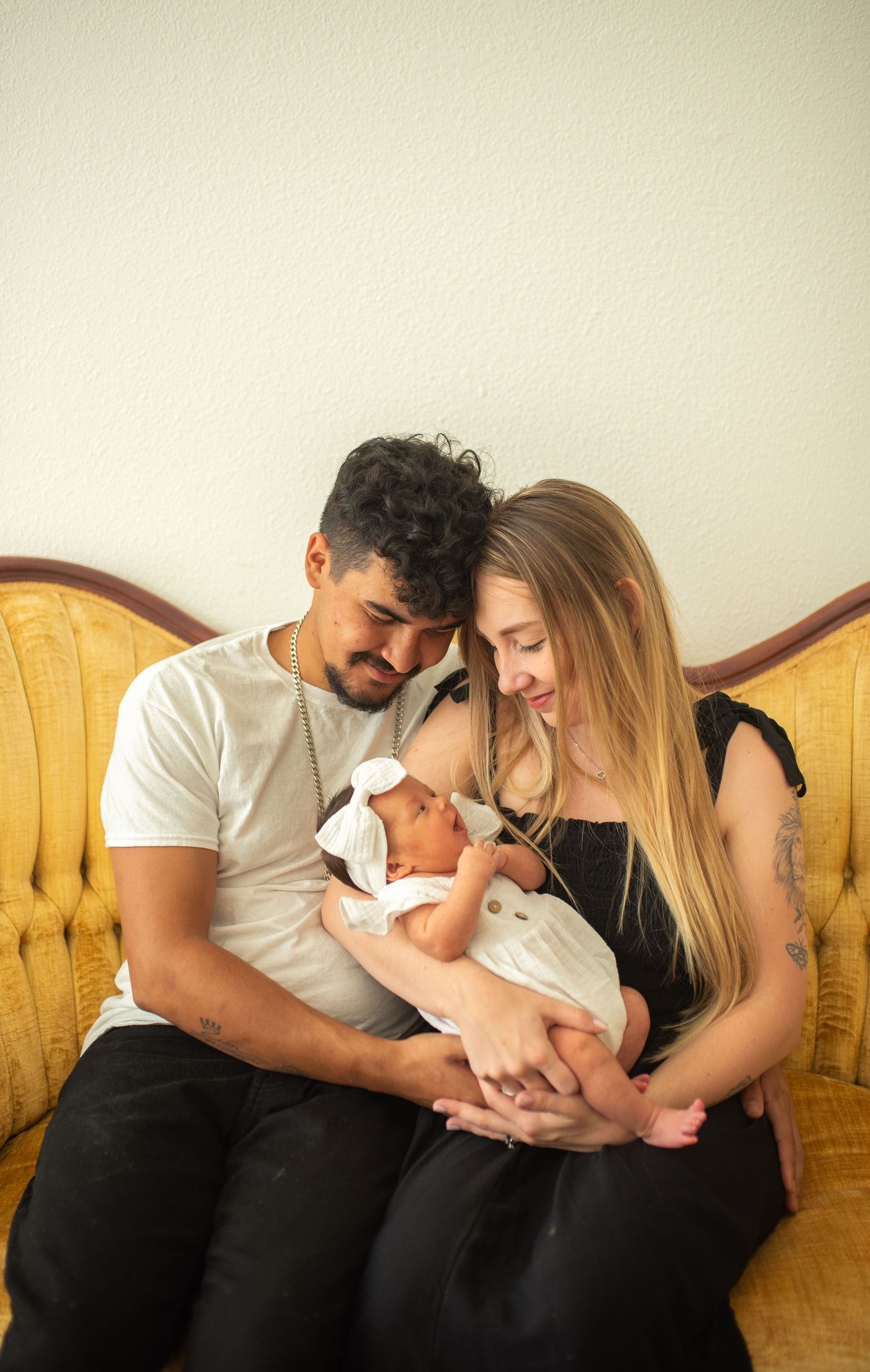 Young couple gazing down tenderly at their sleeping newborn baby on a vintage gold couch.
