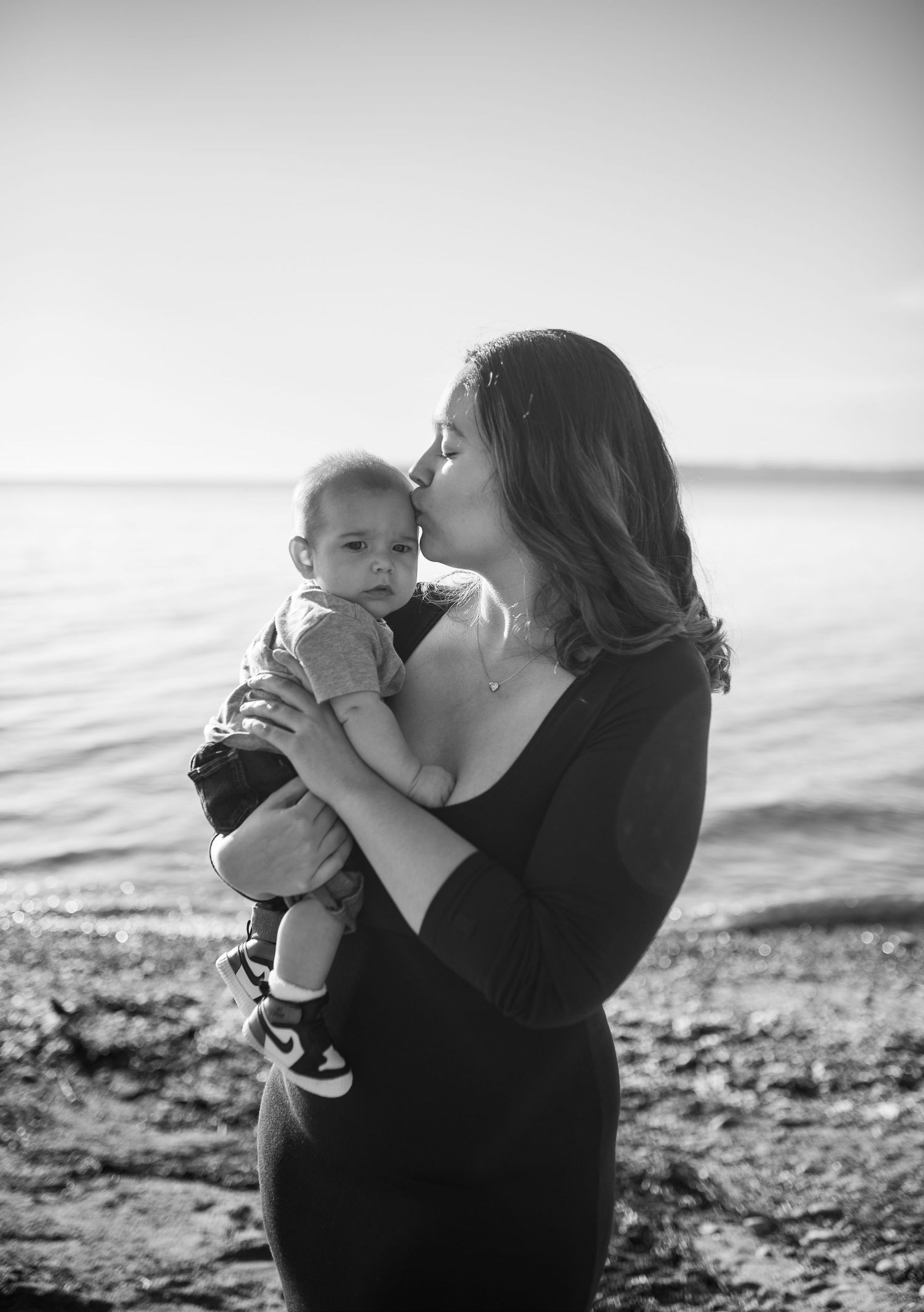 Black and white photo of a mother kissing her baby while standing on a rocky shoreline near calm water.