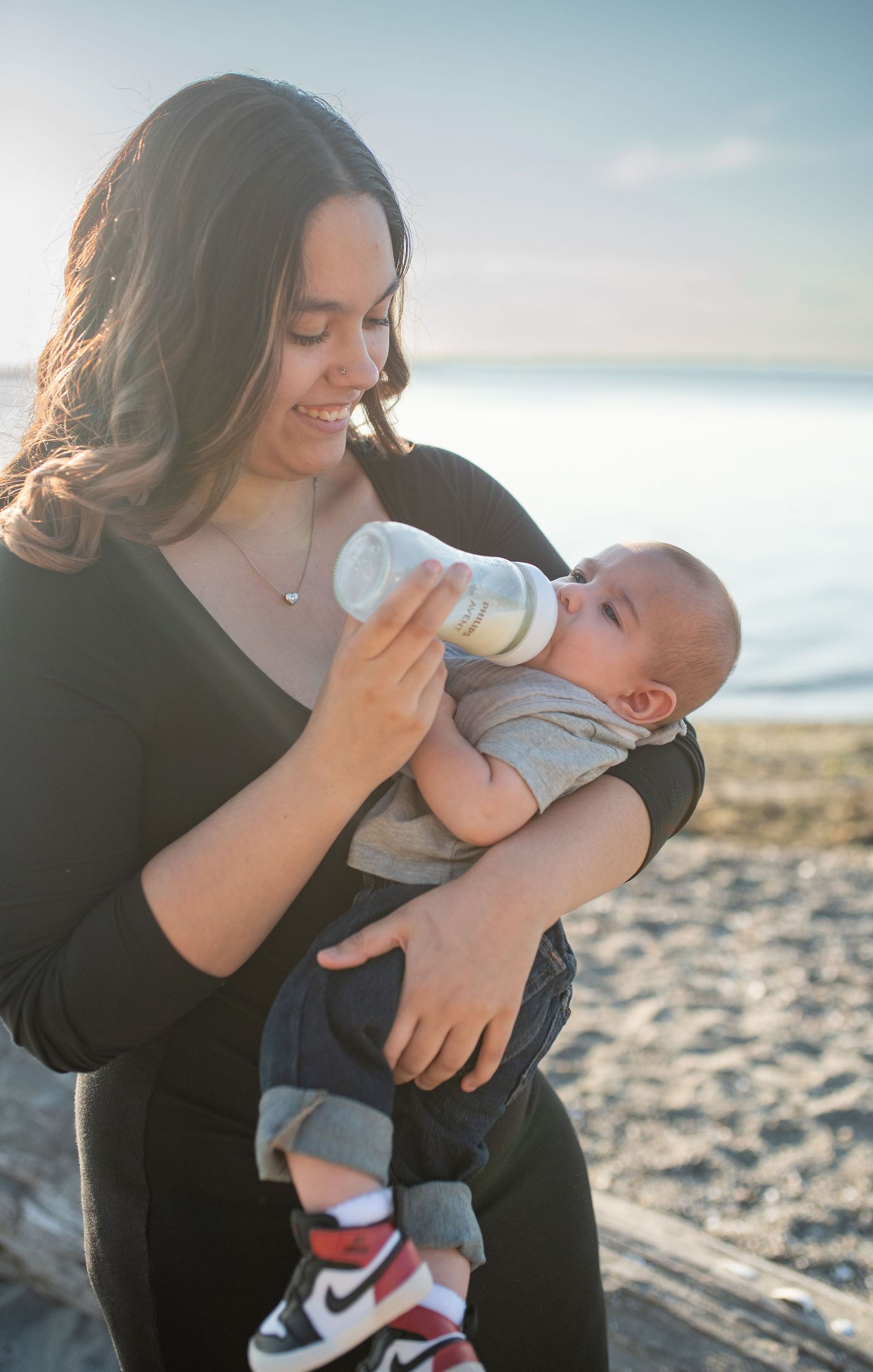Young mother bottle-feeding her newborn baby on a sunny beach, smiling warmly while holding infant in arms.