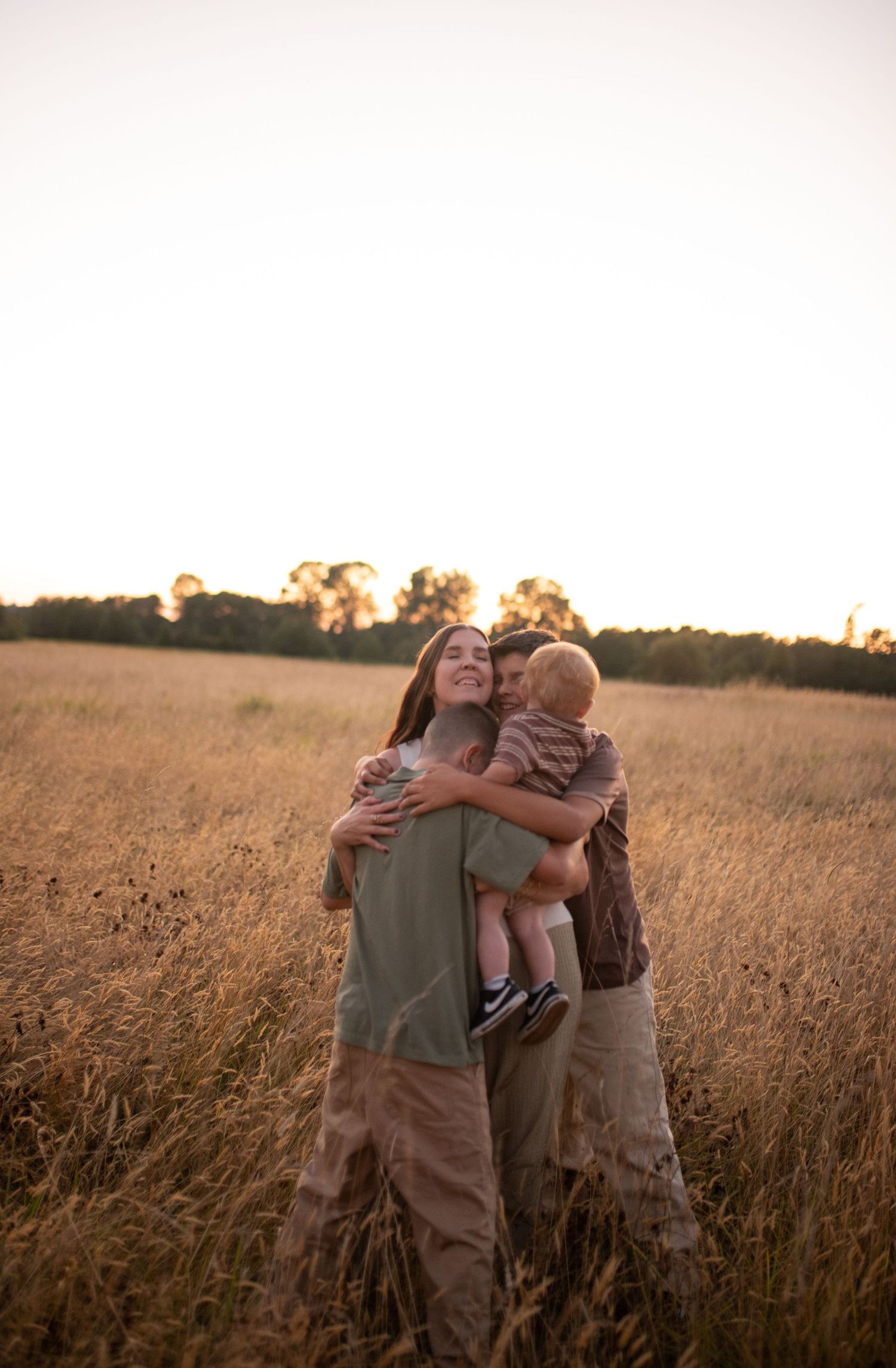 Couple embracing with baby between them in a sunlit golden wheat field at dusk.