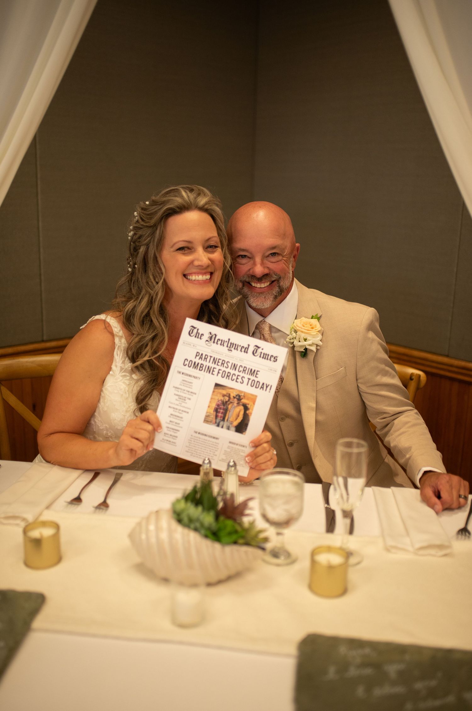 Newlyweds smiling at wedding reception table, bride in white dress holding The Newlywed Times newspaper, groom in tan suit.