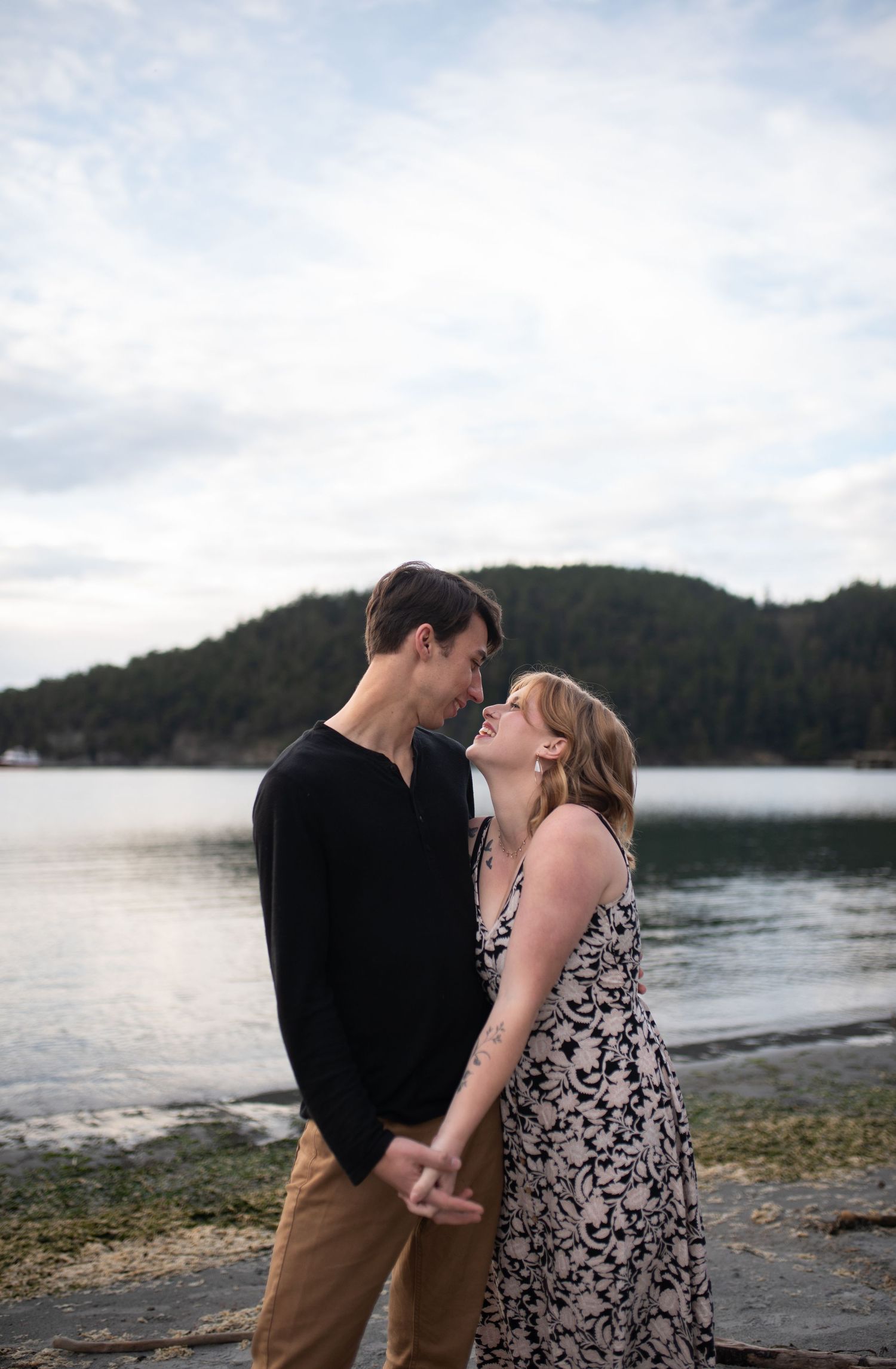 Couple holding hands and laughing together on a pebble beach with a forested island in the background.