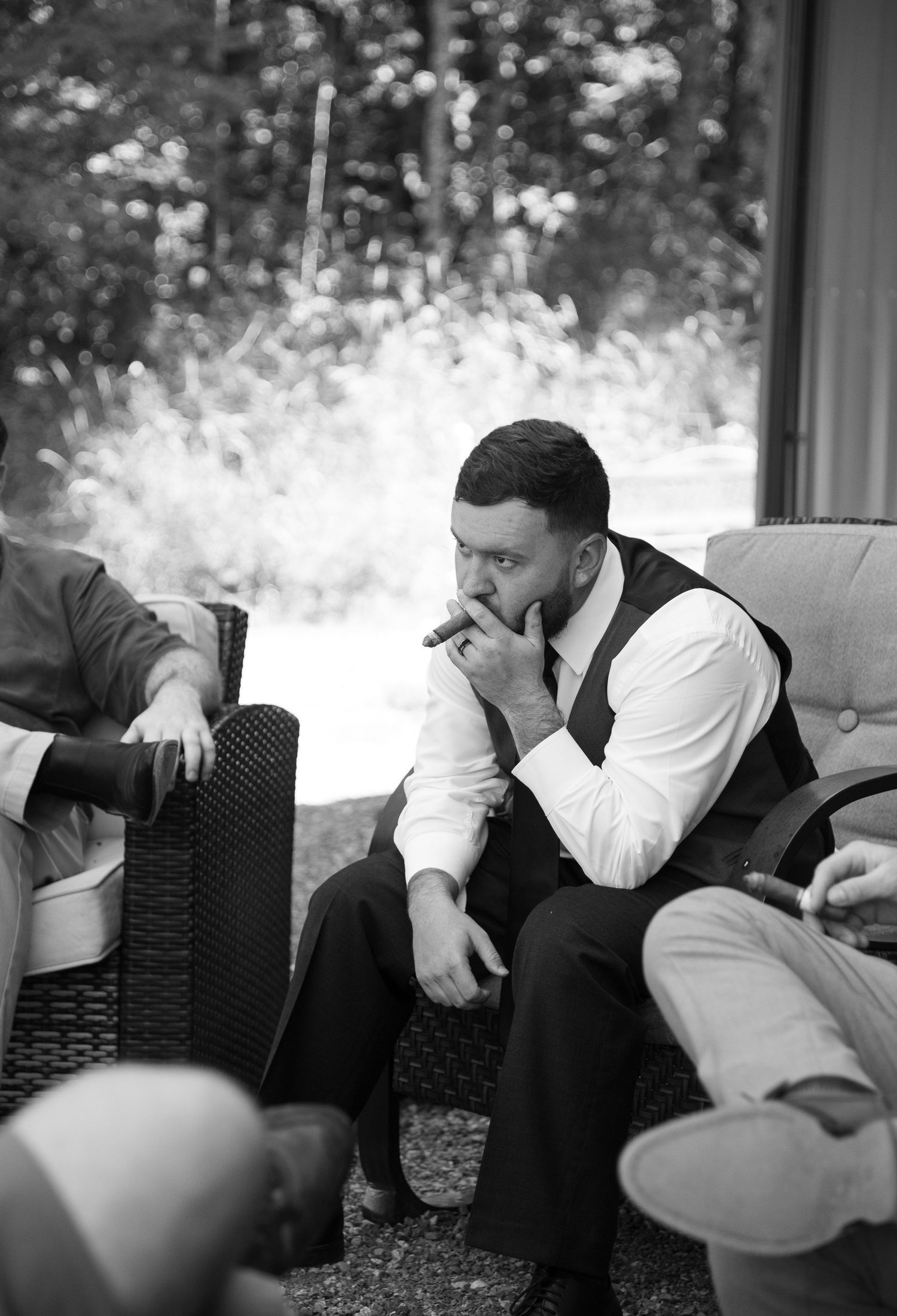Black and white photo of a groom smoking a cigar while relaxing with groomsmen before the wedding ceremony.