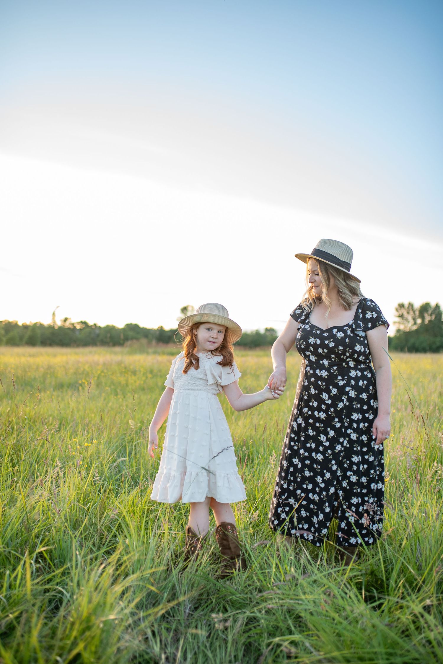Mother and red-haired daughter in straw hats hold hands walking through a sunlit green meadow.