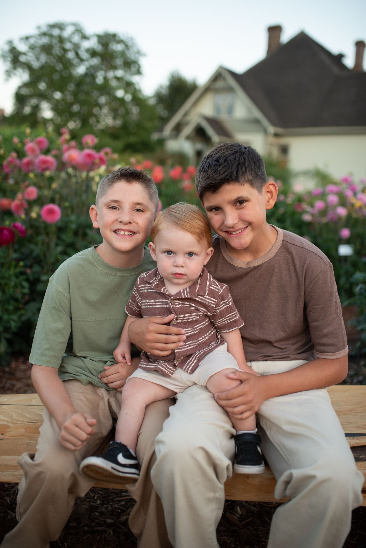 Three boys sitting on a wooden bench in a garden with pink flowers and a house in the background.