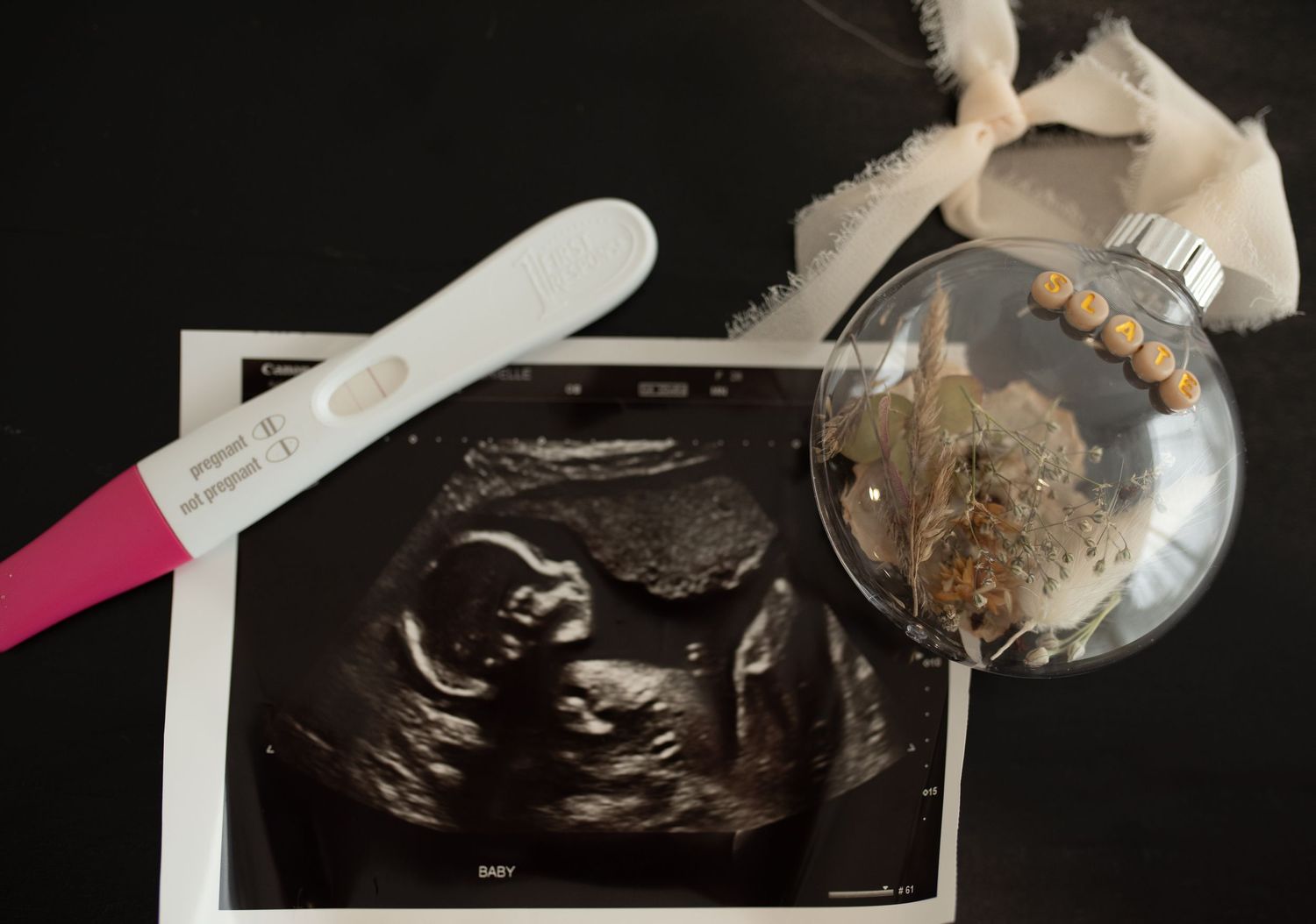 Pregnancy announcement flat lay with ultrasound scan, positive test, and decorative ornament on dark background.