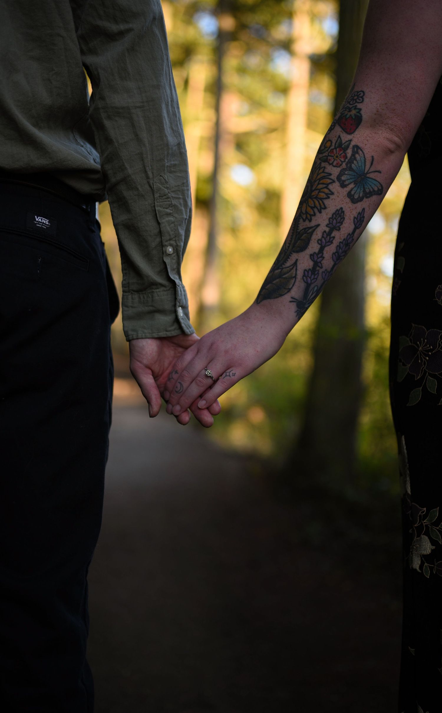 Close-up of couple holding hands revealing intricate butterfly and floral arm tattoos on forest path.