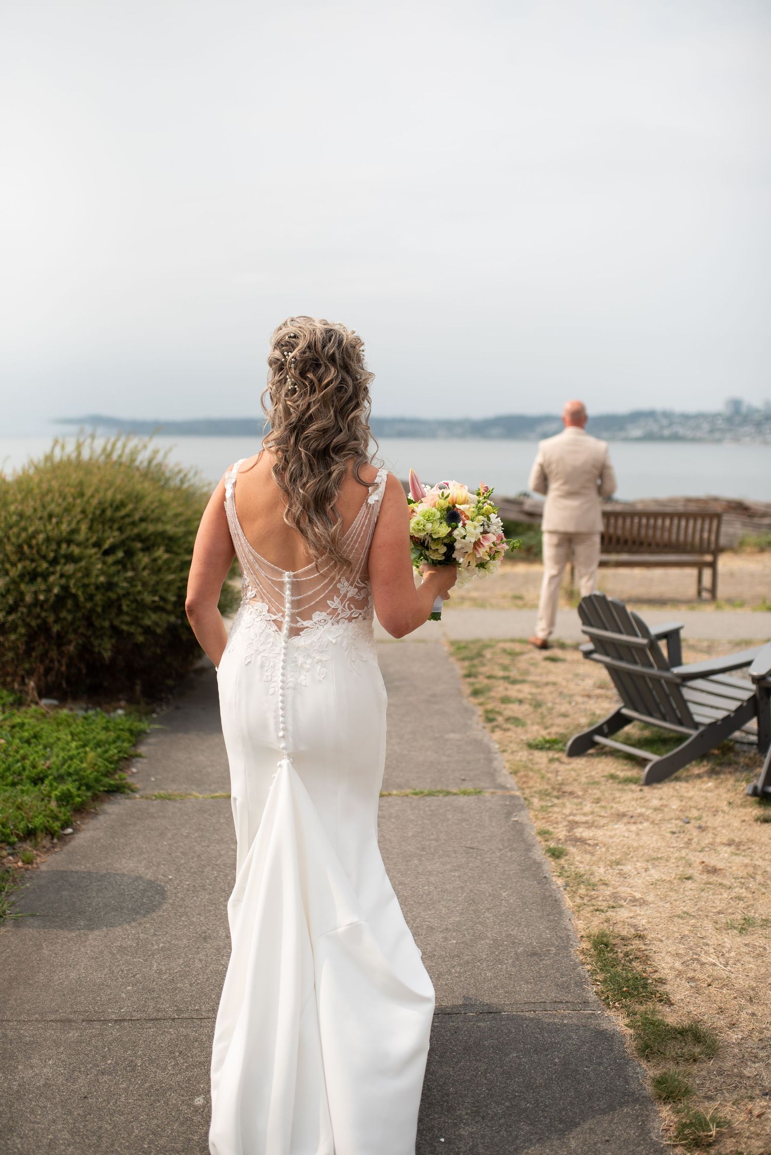 Bride in elegant lace-back wedding gown walks toward groom during first look ceremony by the waterfront.