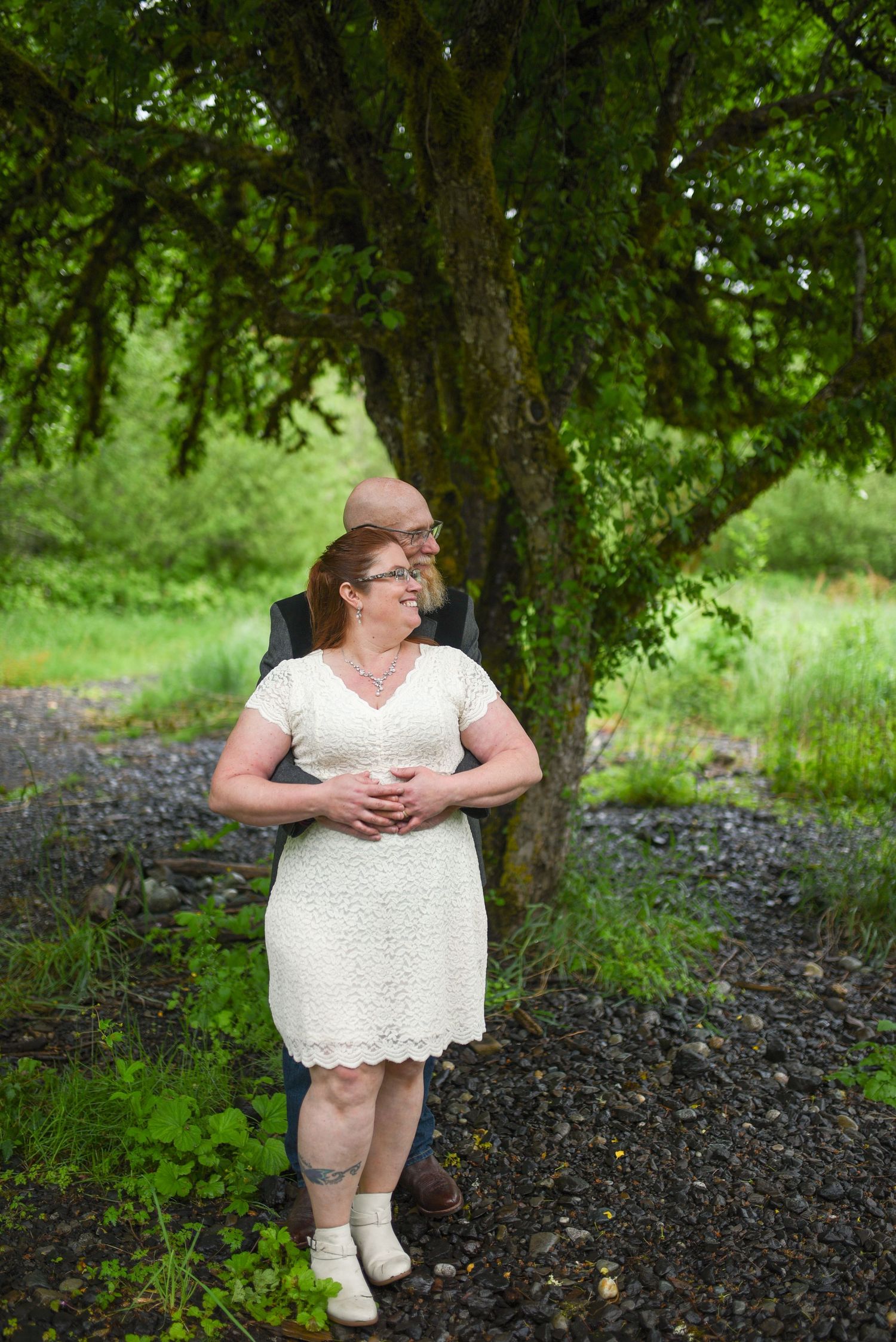 Couple embracing outdoors in wedding attire, bride in white lace dress, groom in suit, under a lush green tree.