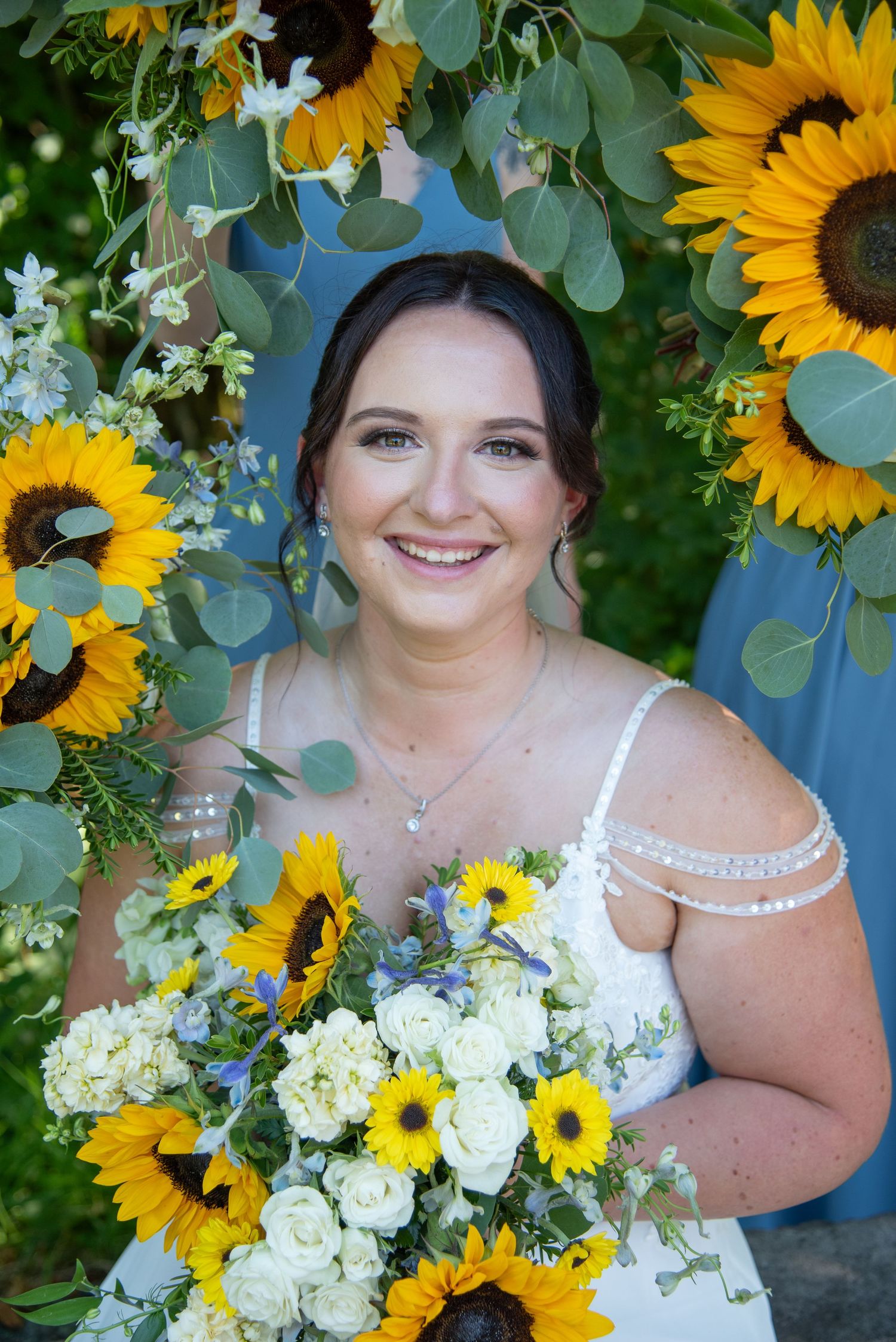 Smiling bride holding a bouquet of sunflowers and white roses, framed by a floral arch of sunflowers and eucalyptus.