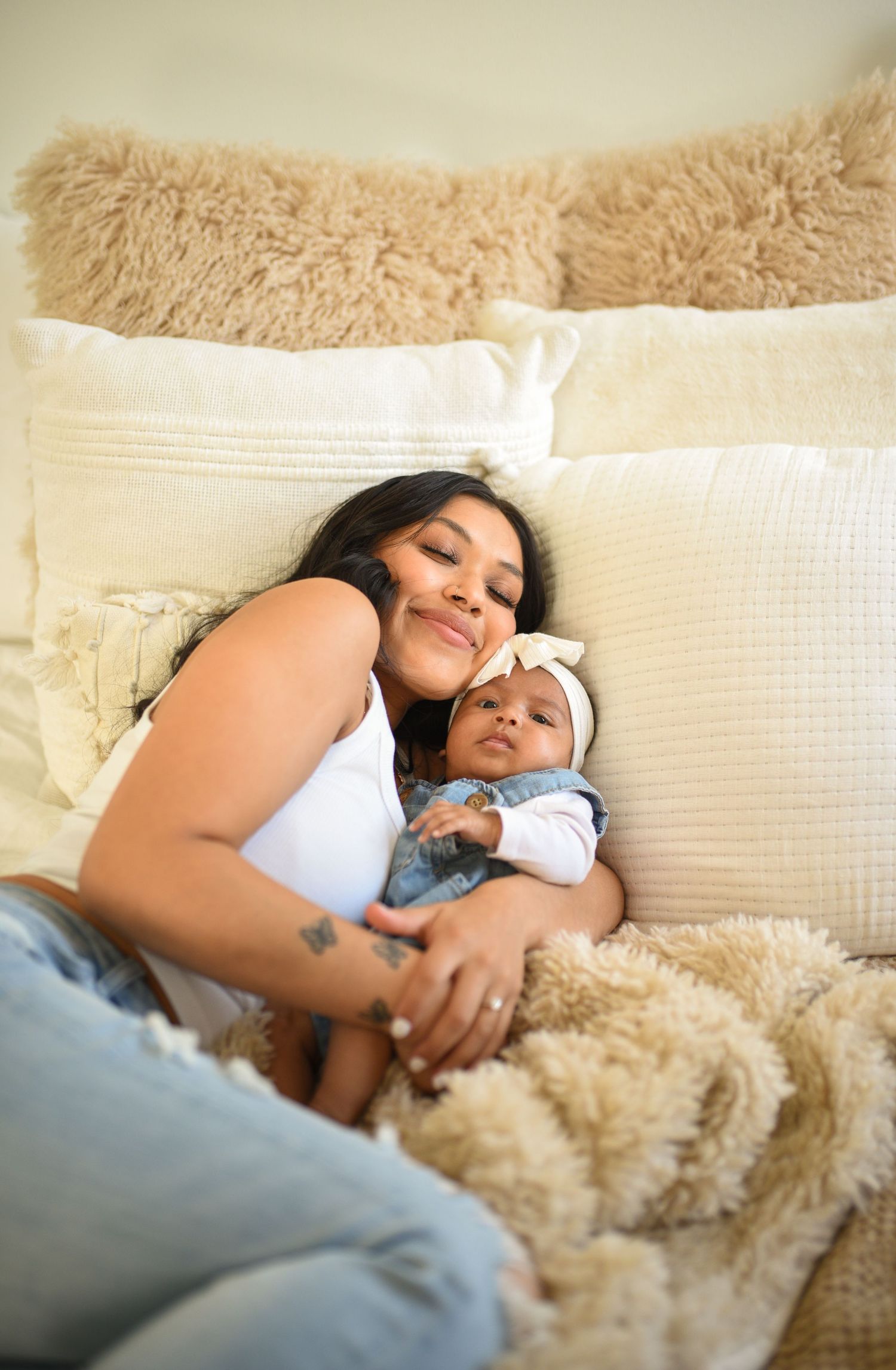 A young mother cuddles her newborn baby on a cozy bed with cream pillows and a fluffy blanket.