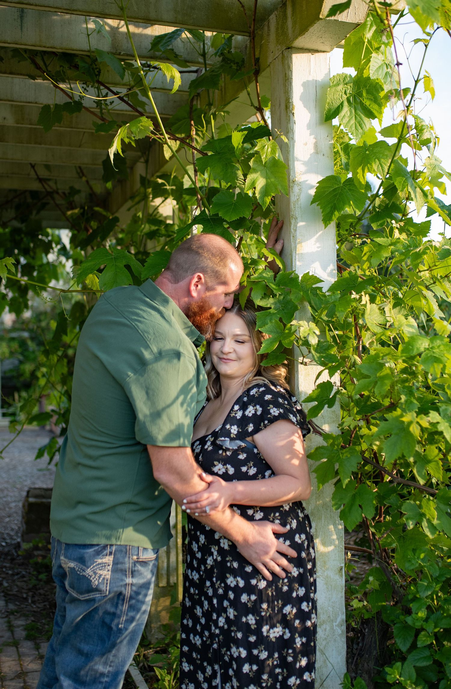 A couple shares a tender moment under a lush green vine-covered pergola, with the man kissing the woman's head as she smiles.