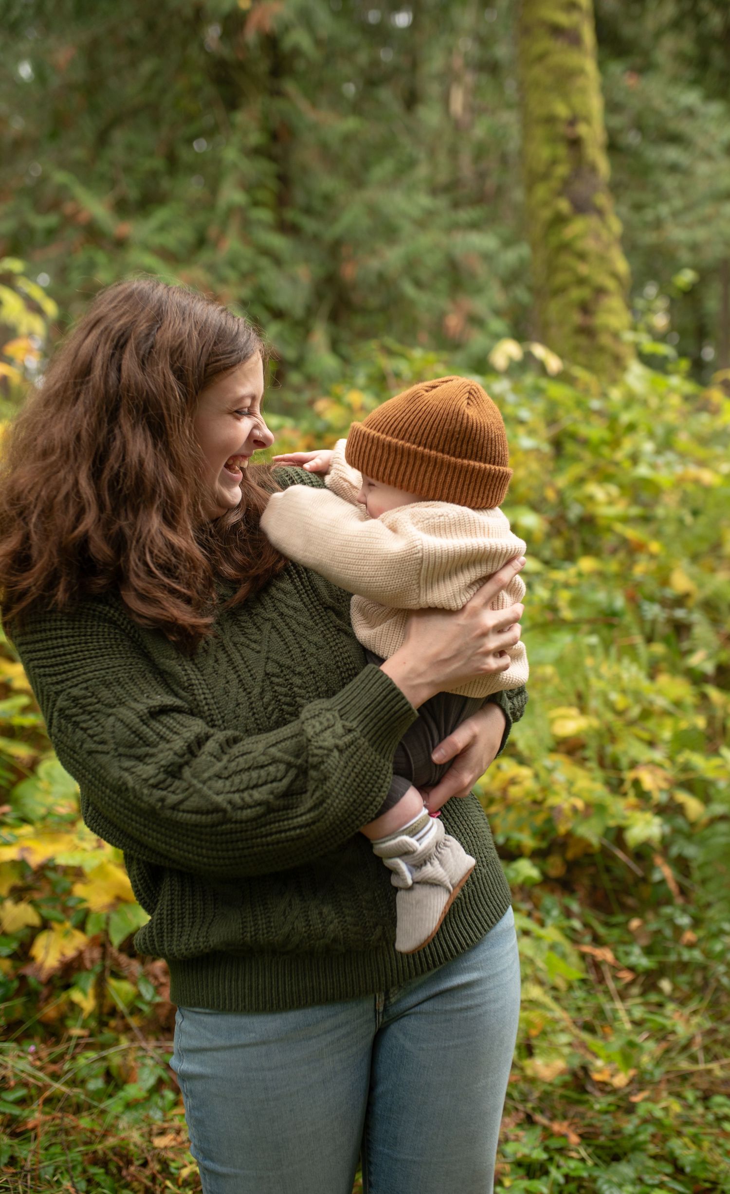 A smiling woman in a green sweater holds a baby in a beige outfit and brown knit hat in a lush forest.