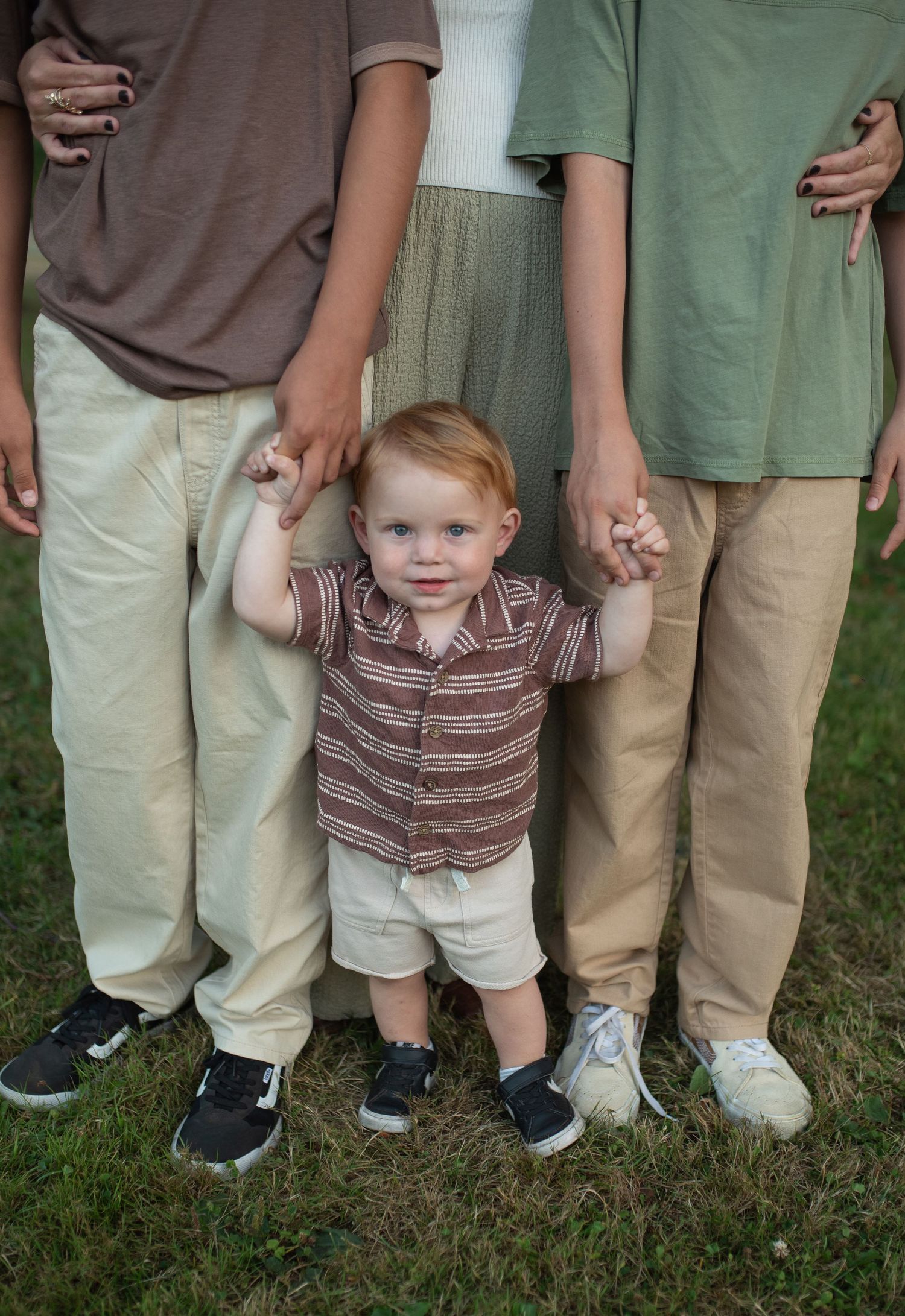 Toddler boy in striped shirt holding hands with two older siblings standing on green grass.