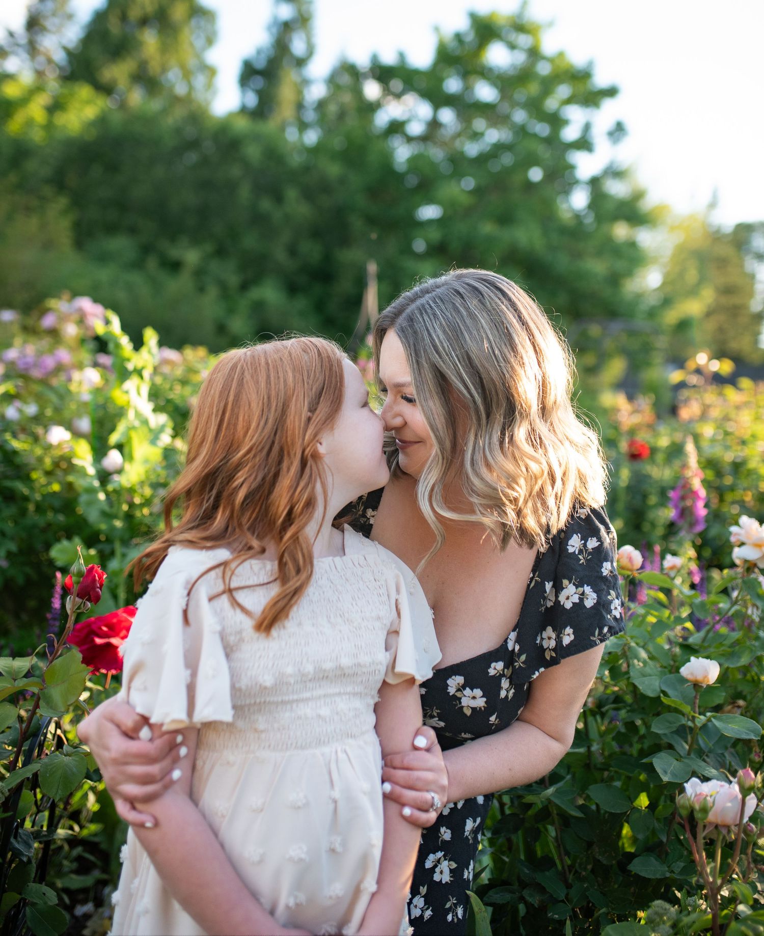Red-haired girl and blonde woman share a tender nose-to-nose moment surrounded by colorful garden roses.