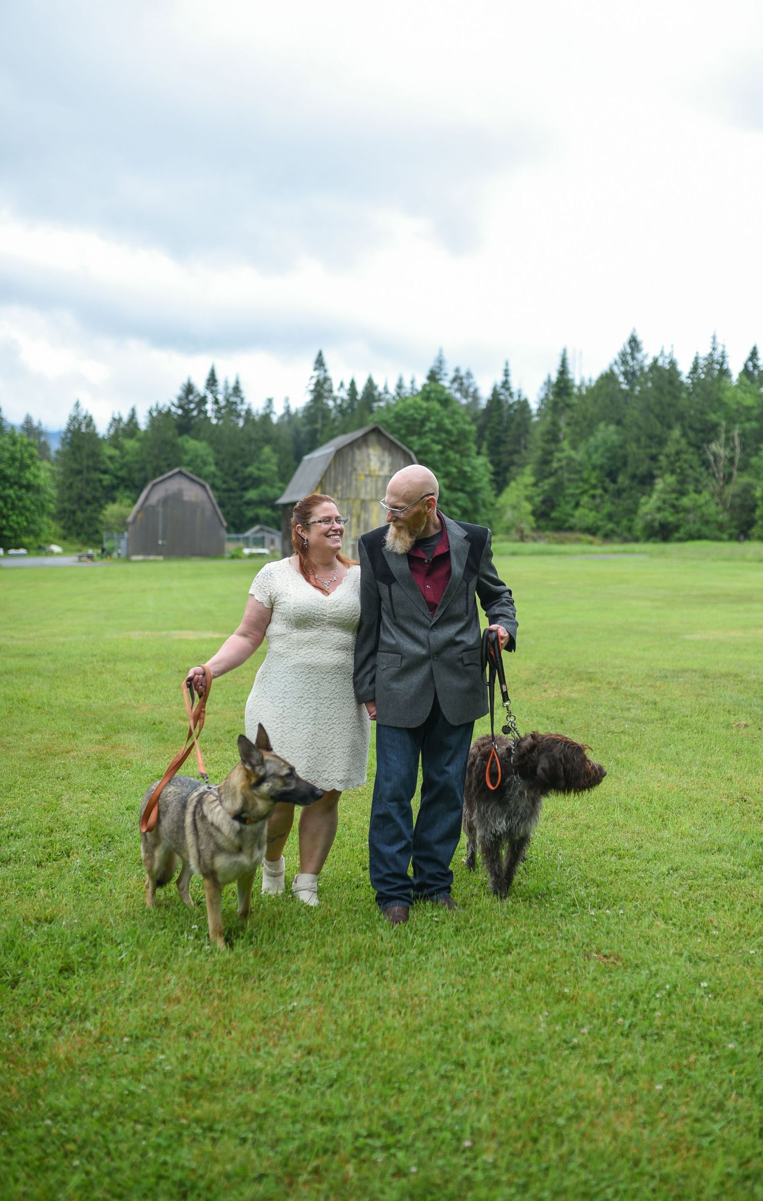 Happy couple in wedding attire walking dogs on green farm lawn with rustic barn and trees in background.