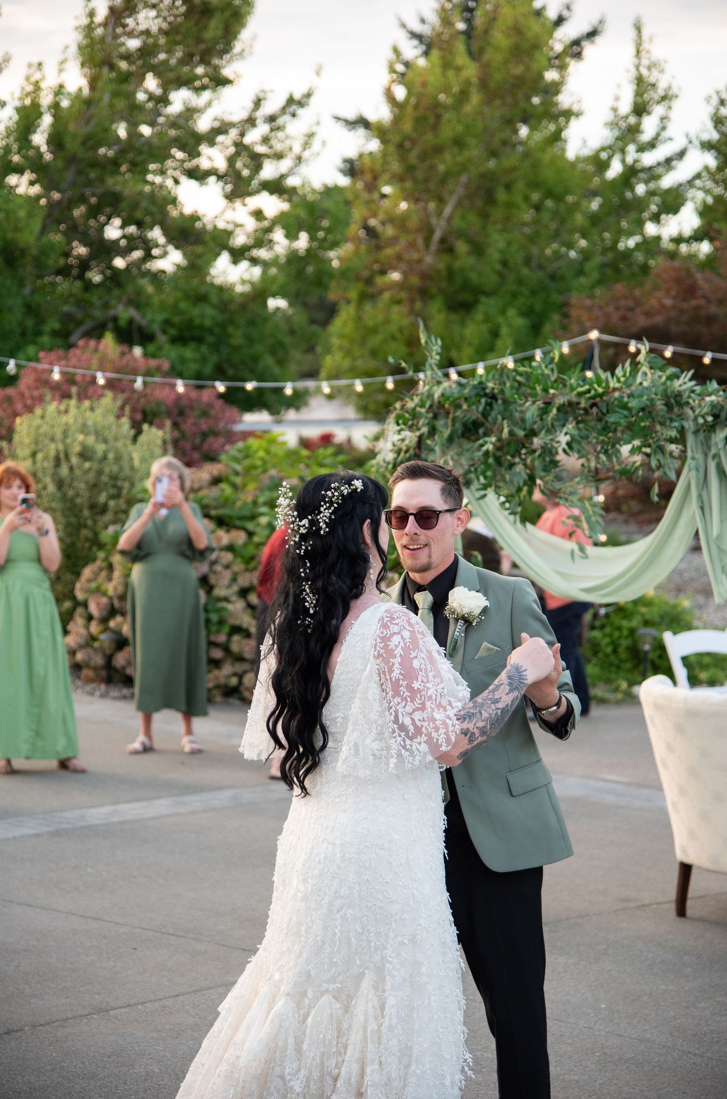 Bride and groom share first dance outdoors at garden wedding reception with green floral arch and string lights.