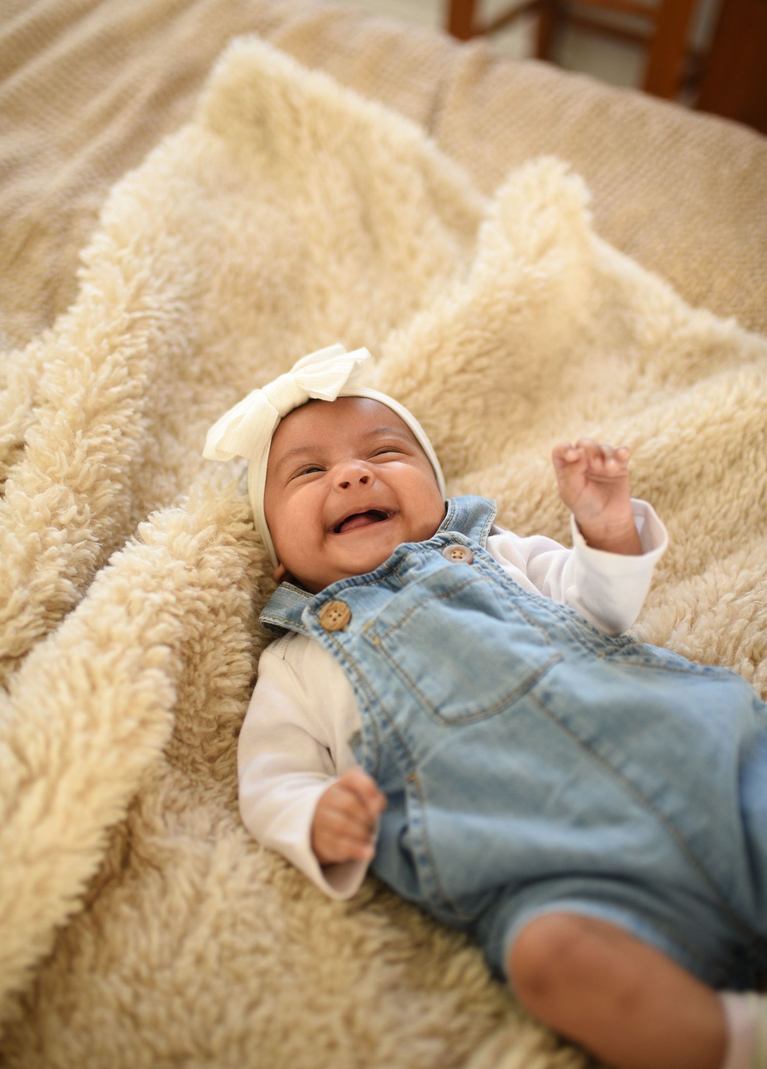 Smiling baby in denim overalls and white bow headband lying on a fluffy cream blanket.
