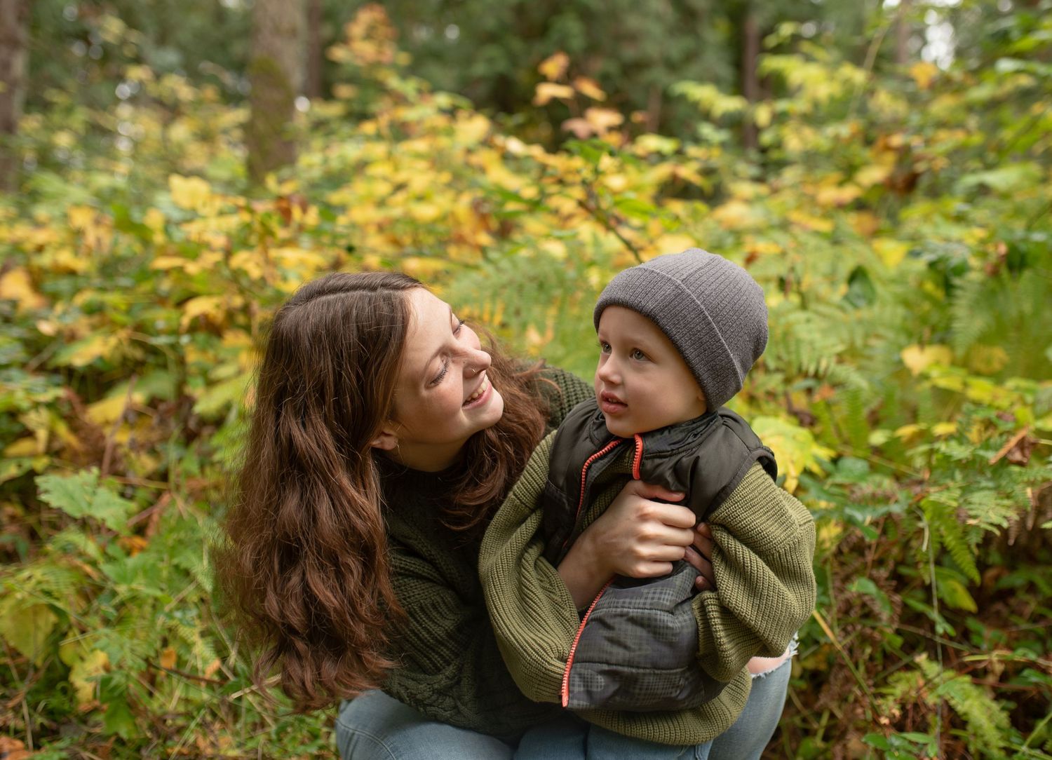 A mother holds her young child in an autumn forest, both smiling warmly among colorful fall foliage.