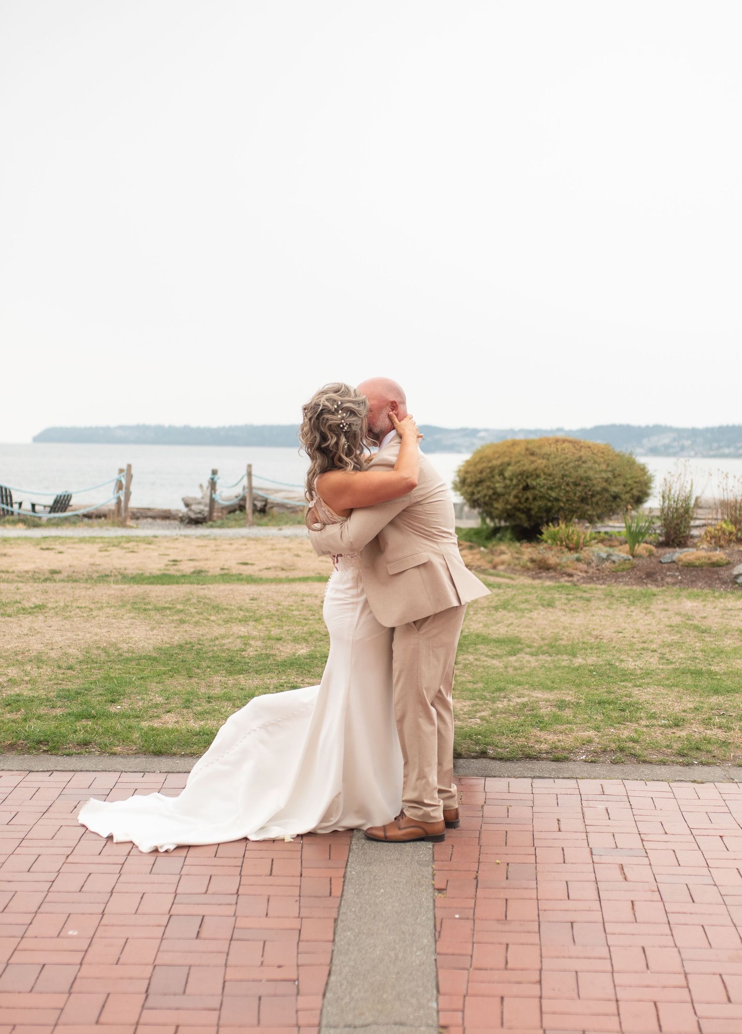 Bride and groom embracing on brick path with waterfront backdrop on their wedding day.