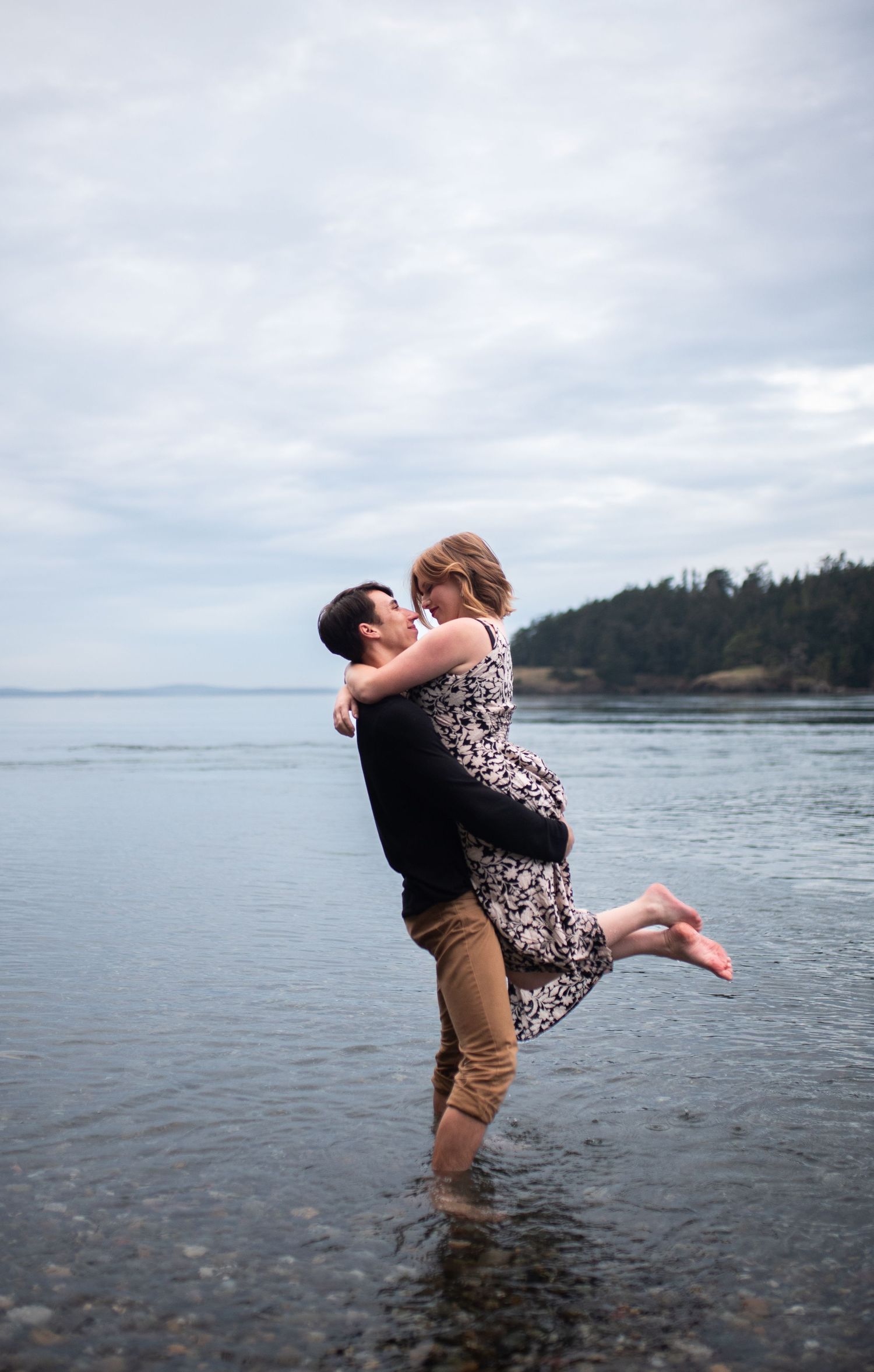 Romantic couple embrace as man lifts woman at the ocean shoreline under cloudy skies.