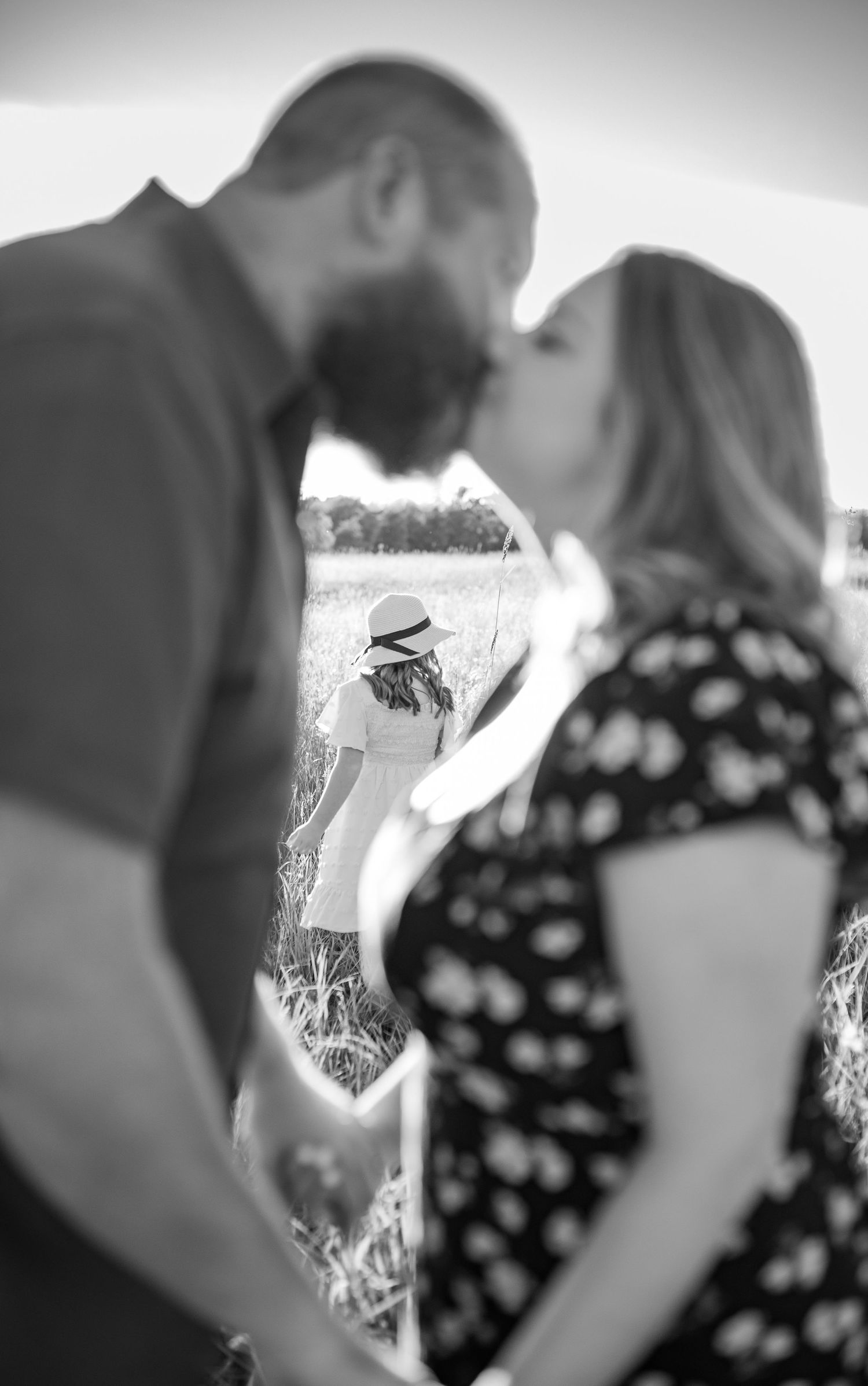 Black and white photo of a couple kissing in a field with a child wearing a hat standing in the background.