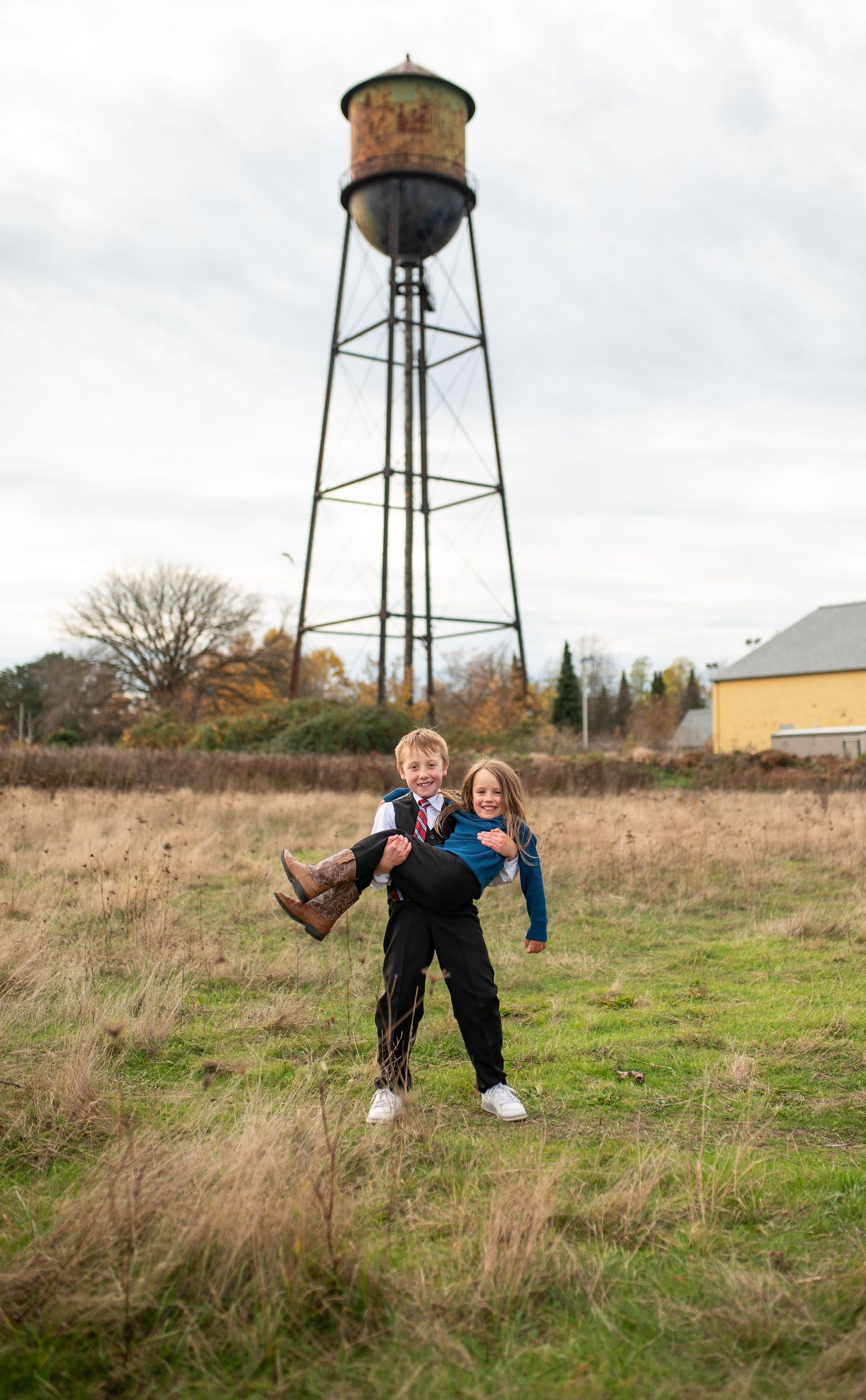Two children laugh as a boy gives a girl a piggyback ride in a field with a water tower behind.