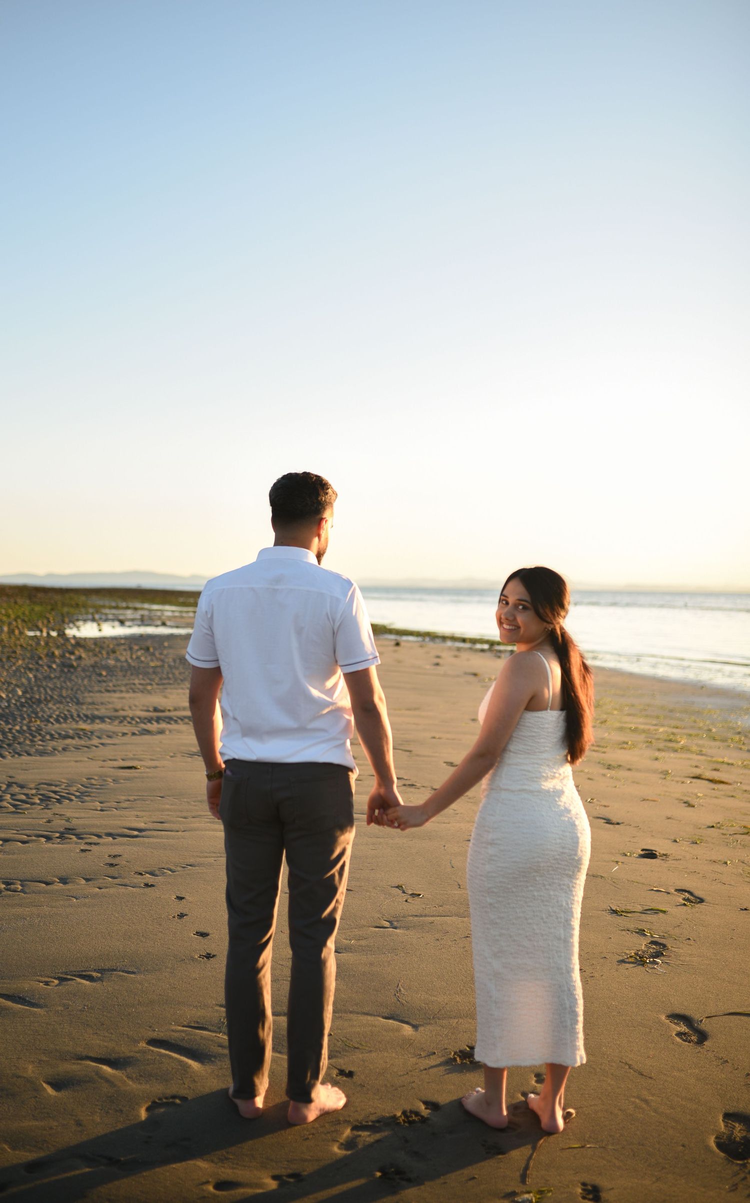Couple holding hands on a sandy beach at sunset, woman in white dress looking back, man in white shirt and gray pants.