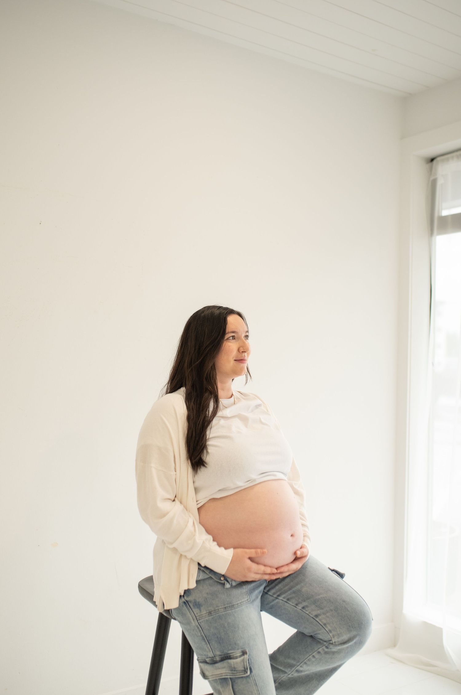 Pregnant woman in white cardigan and jeans sitting on stool cradling her bare baby bump in bright white studio.