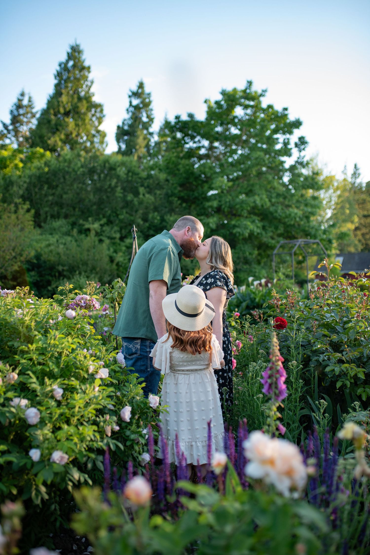 Couple kissing in flower garden while young daughter watches in straw hat.