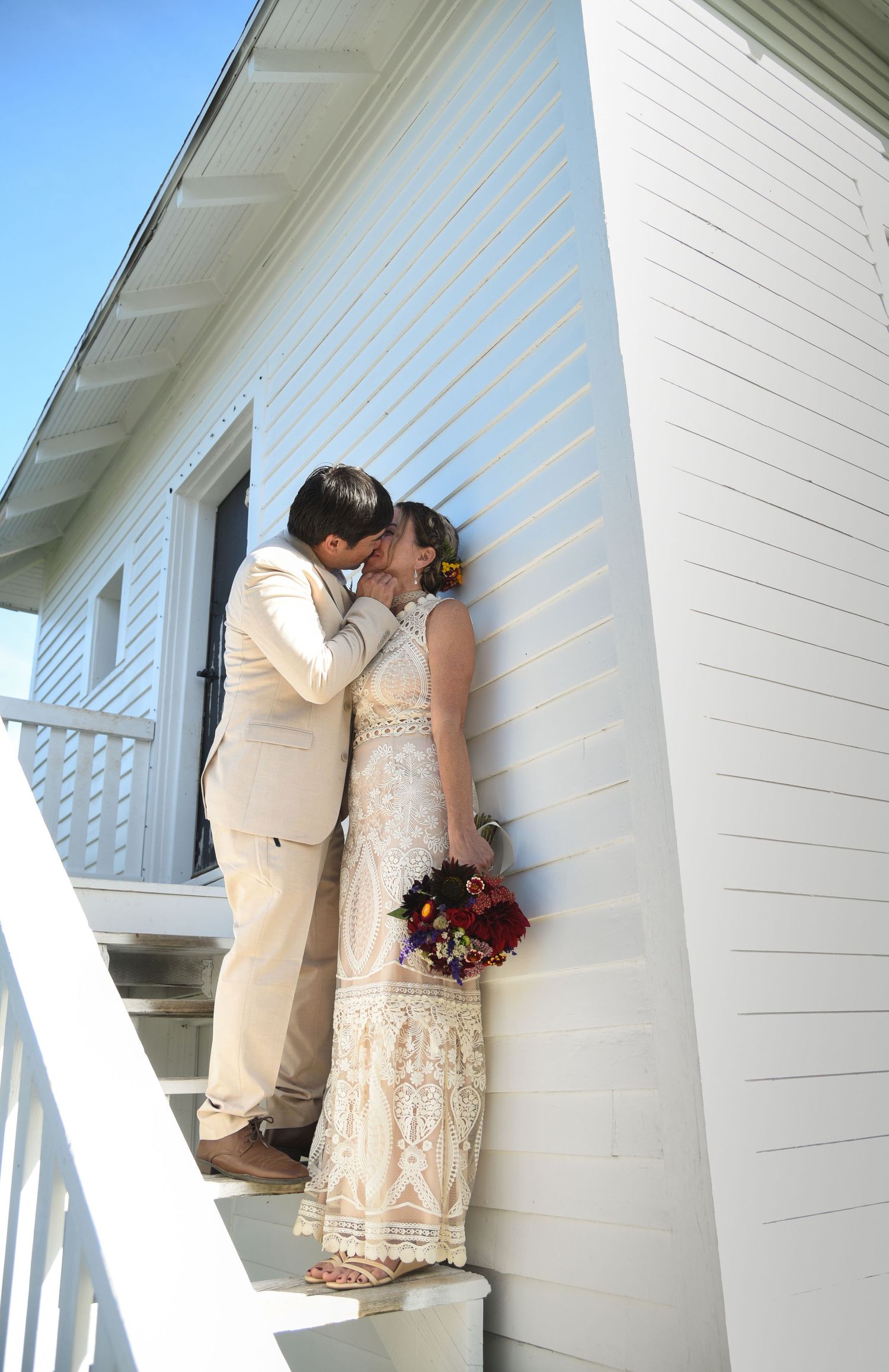 Bride and groom sharing a kiss on white building stairs, bride holding colorful bouquet.