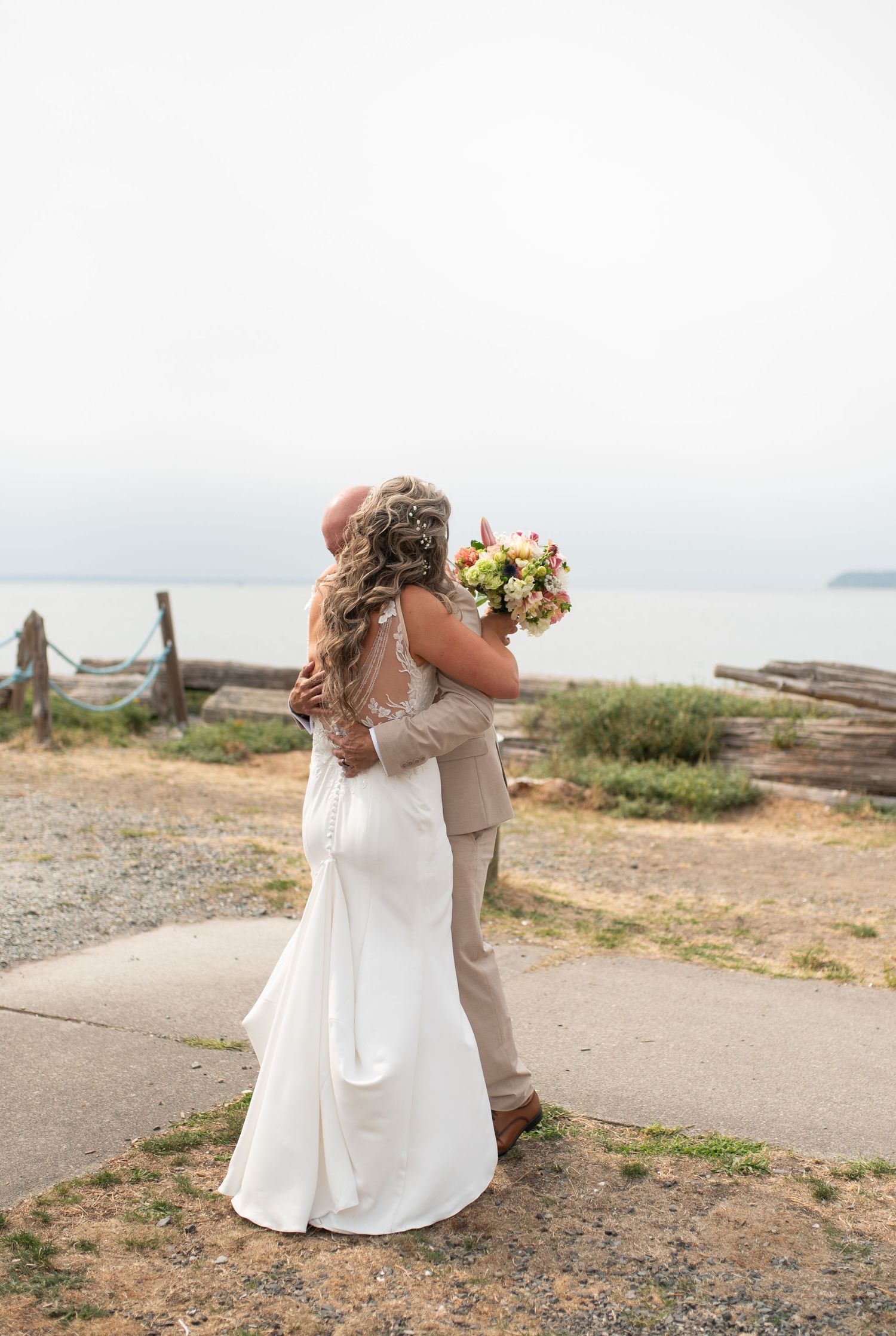Bride and groom embracing on a beachside path, bride holding colorful bouquet in white gown, groom in tan suit.