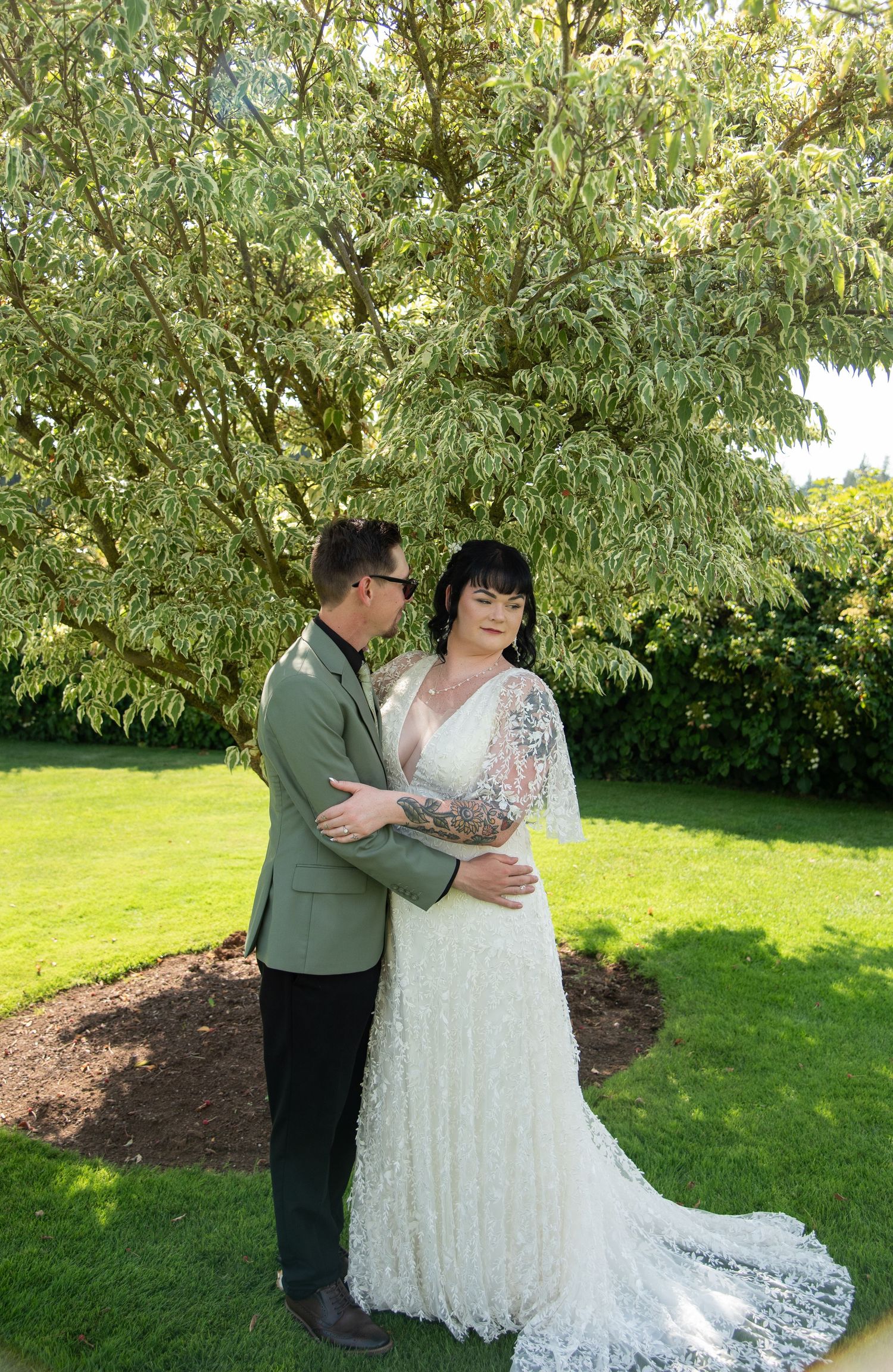 Bride and groom embrace in lush green garden beneath a large leafy tree on wedding day.