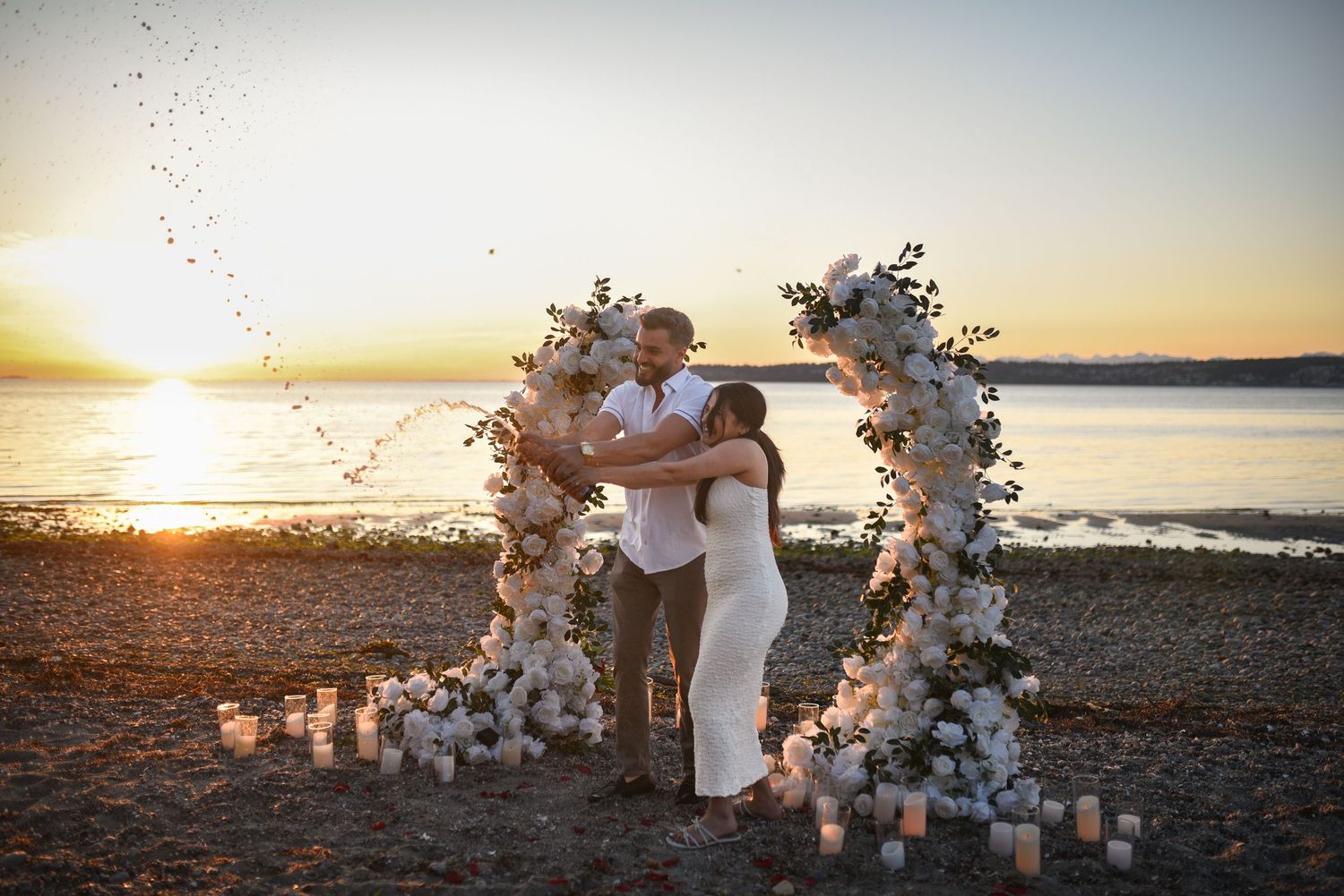 Couple embraces under floral arch on beach at sunset with candles and white roses for romantic wedding ceremony.