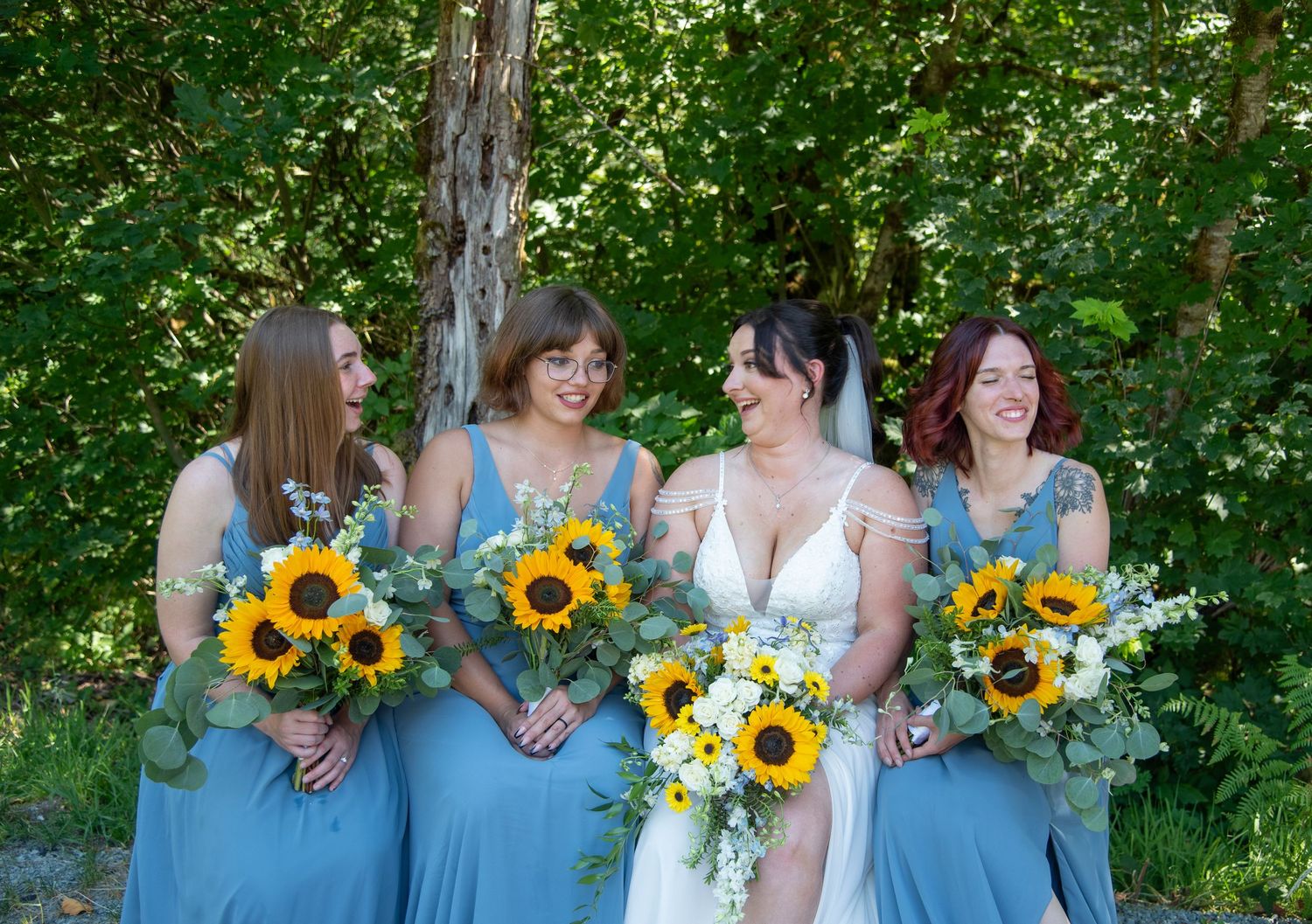 A bride and three bridesmaids in blue dresses laugh together holding sunflower bouquets outdoors.