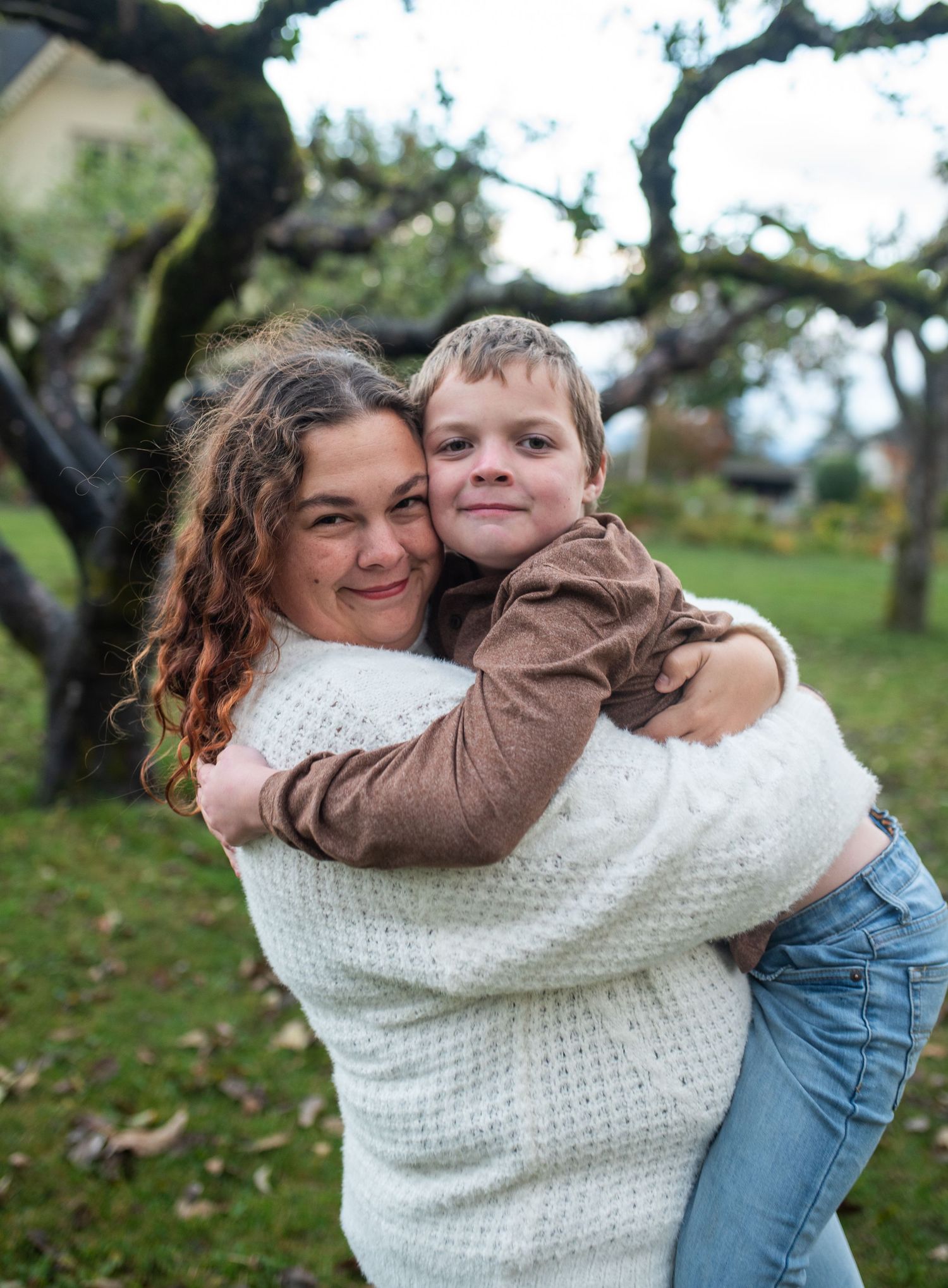 Smiling mother hugging her young son outdoors under mossy trees in autumn.