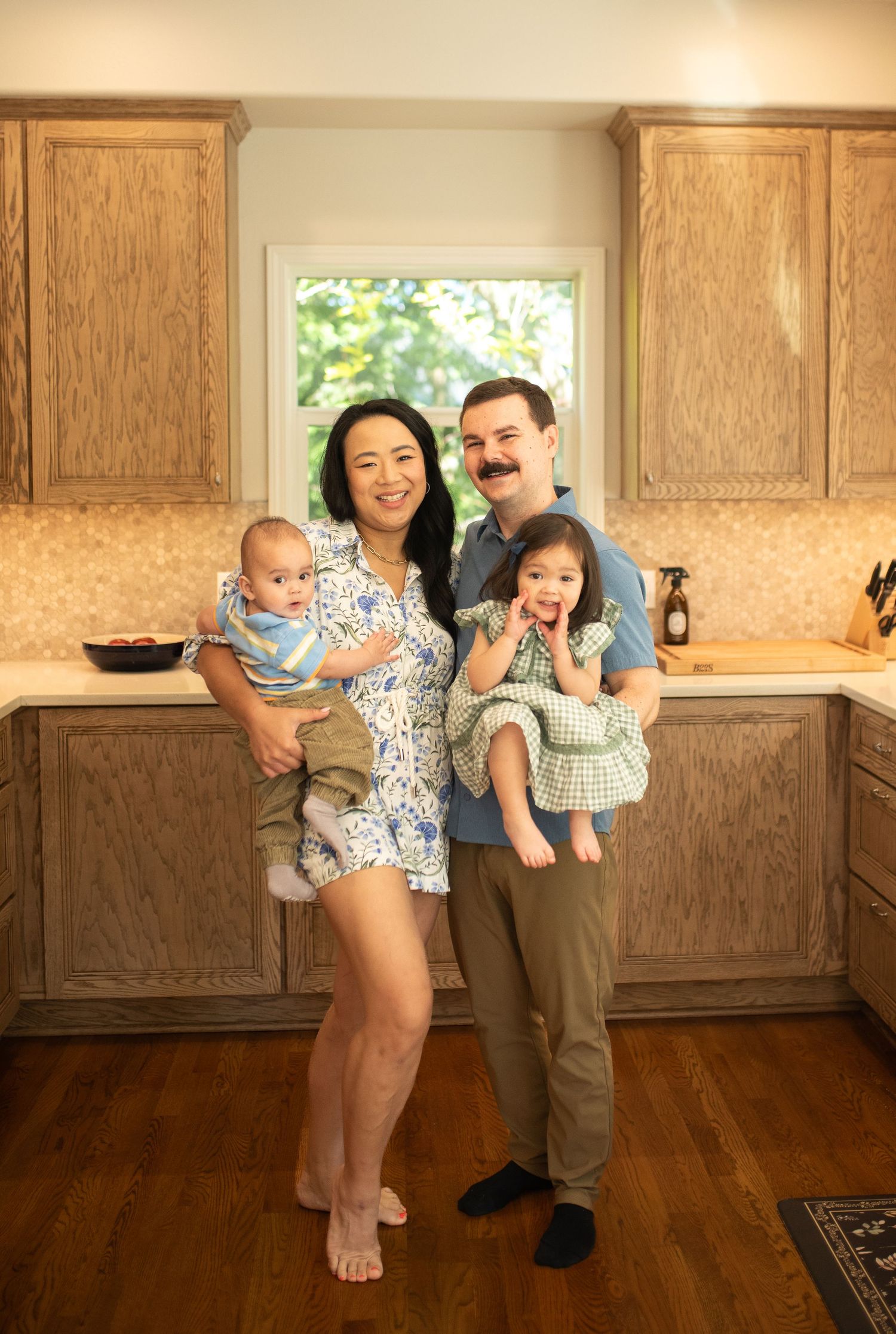 Smiling family portrait in a cozy kitchen, parents holding their baby boy and toddler daughter in matching outfits.