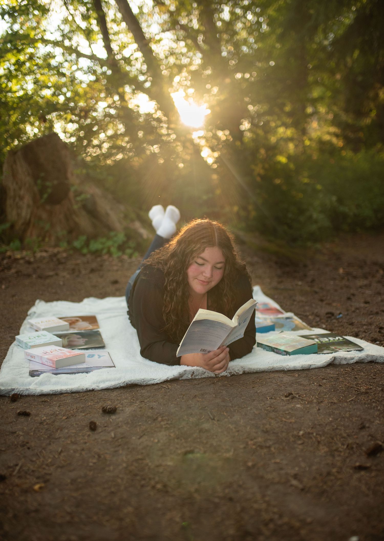 Teen girl reads book lying on blanket in sunlit forest clearing.