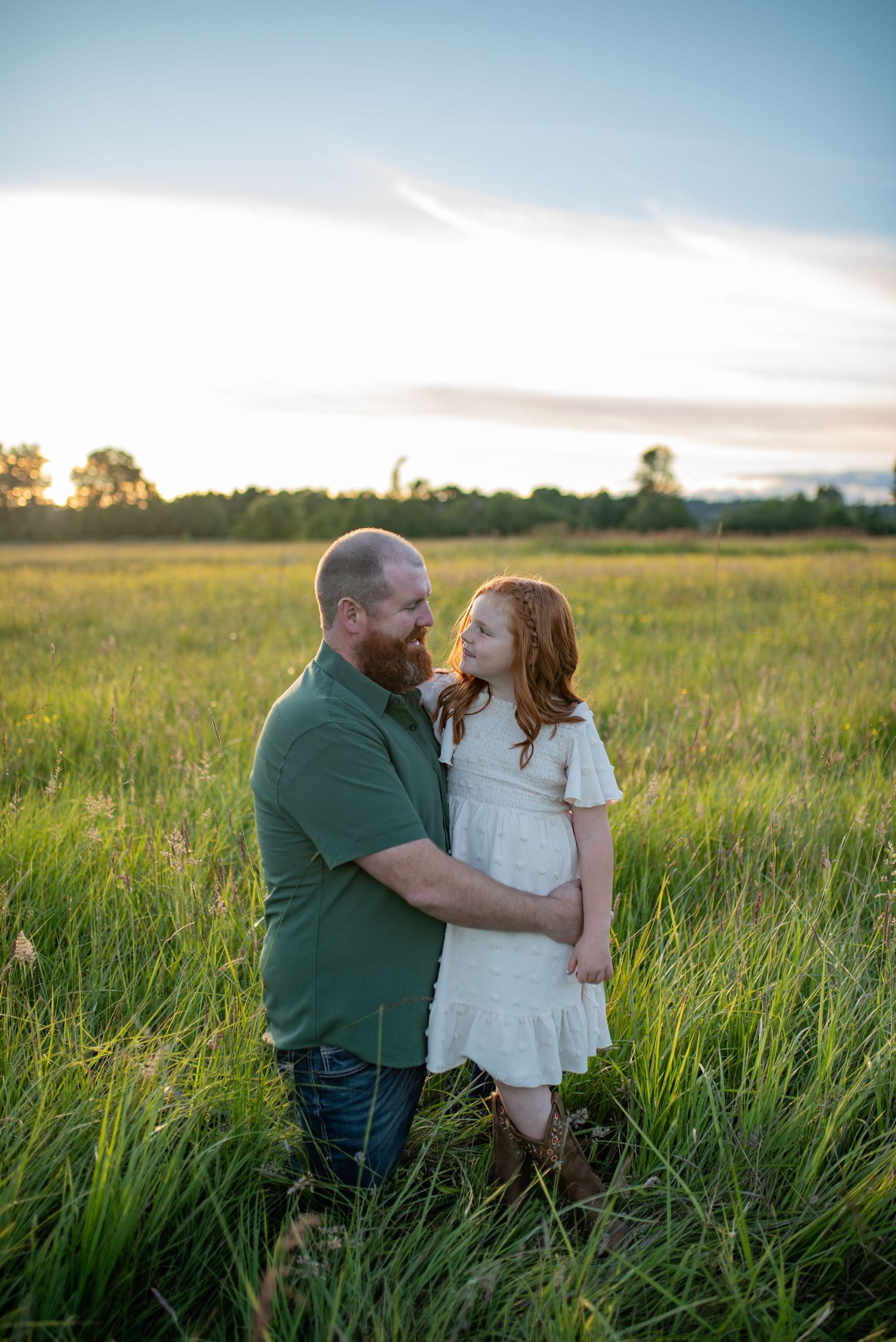 Bearded father and red-haired daughter share a tender moment in a lush green meadow at sunset.