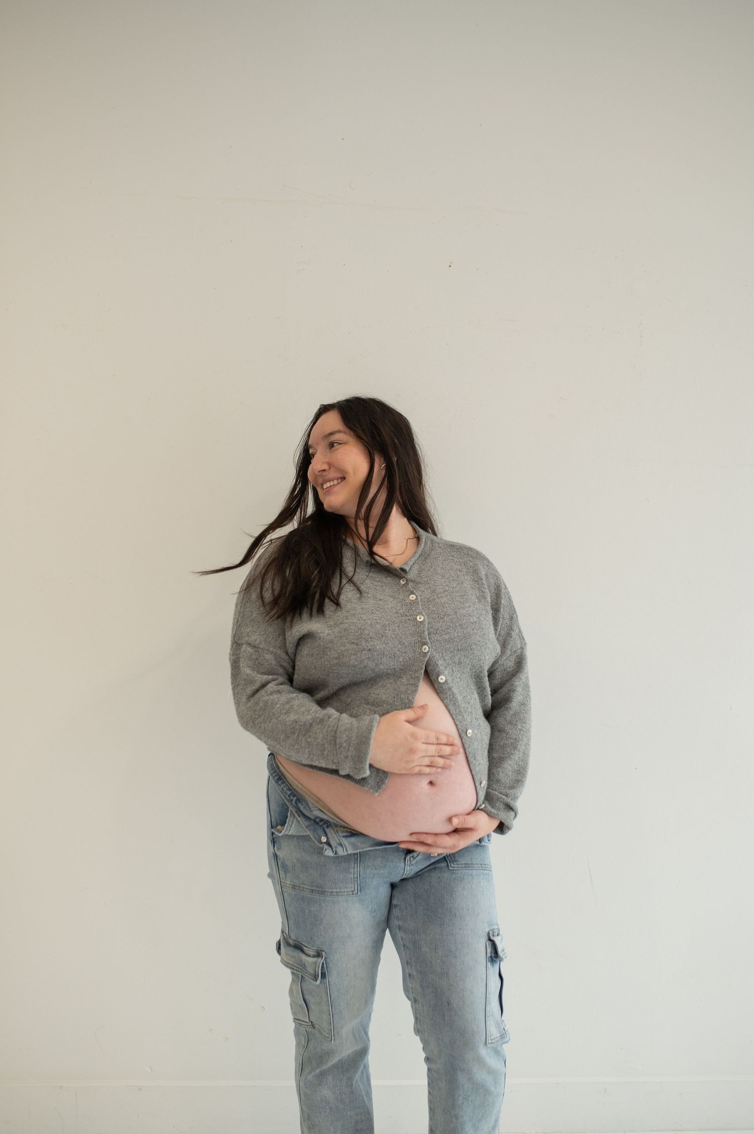 Smiling pregnant woman in gray cardigan and cargo jeans cradling her baby bump against a clean white studio wall.