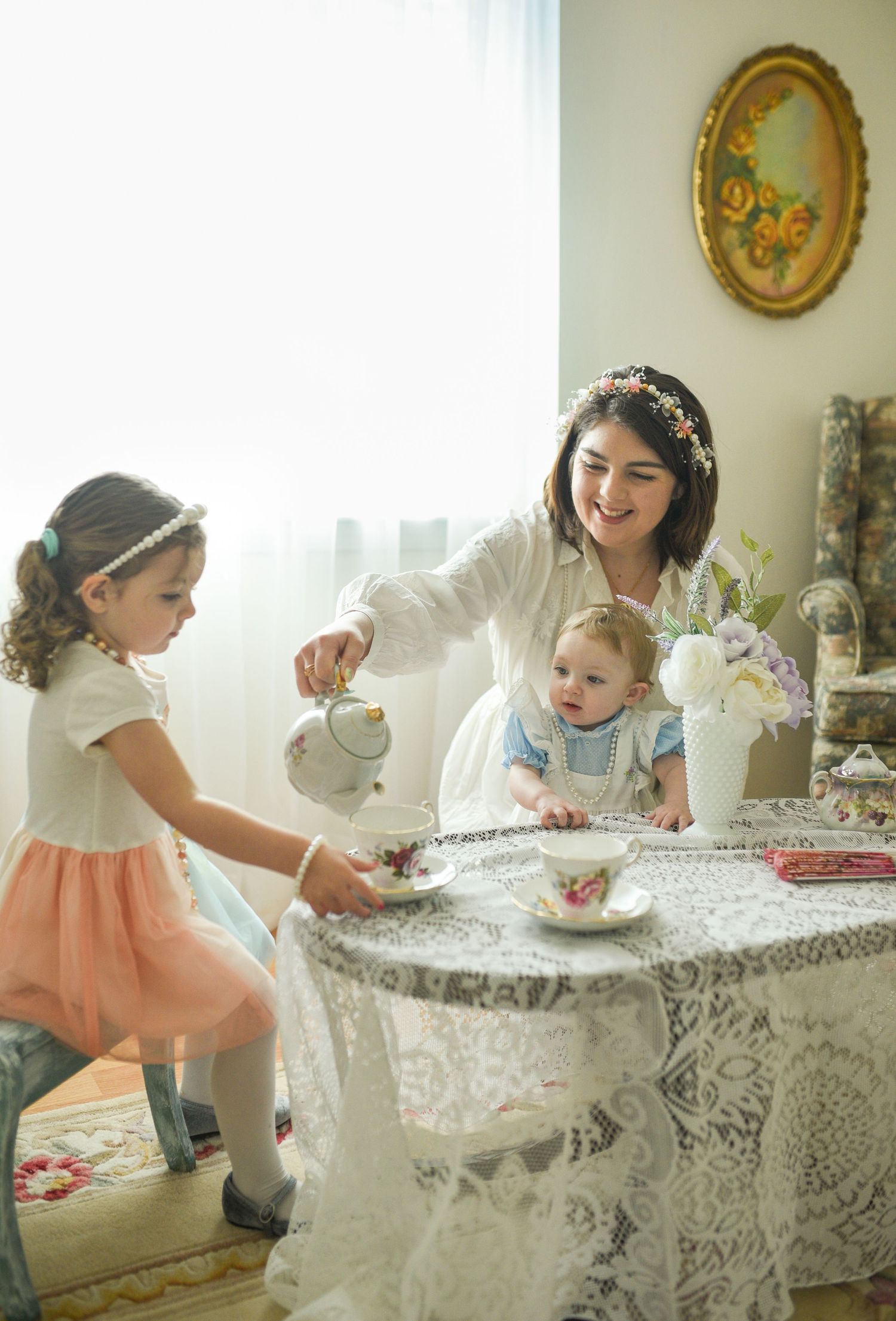 A mother in a floral crown pours tea for two young daughters at a lace-covered table indoors.