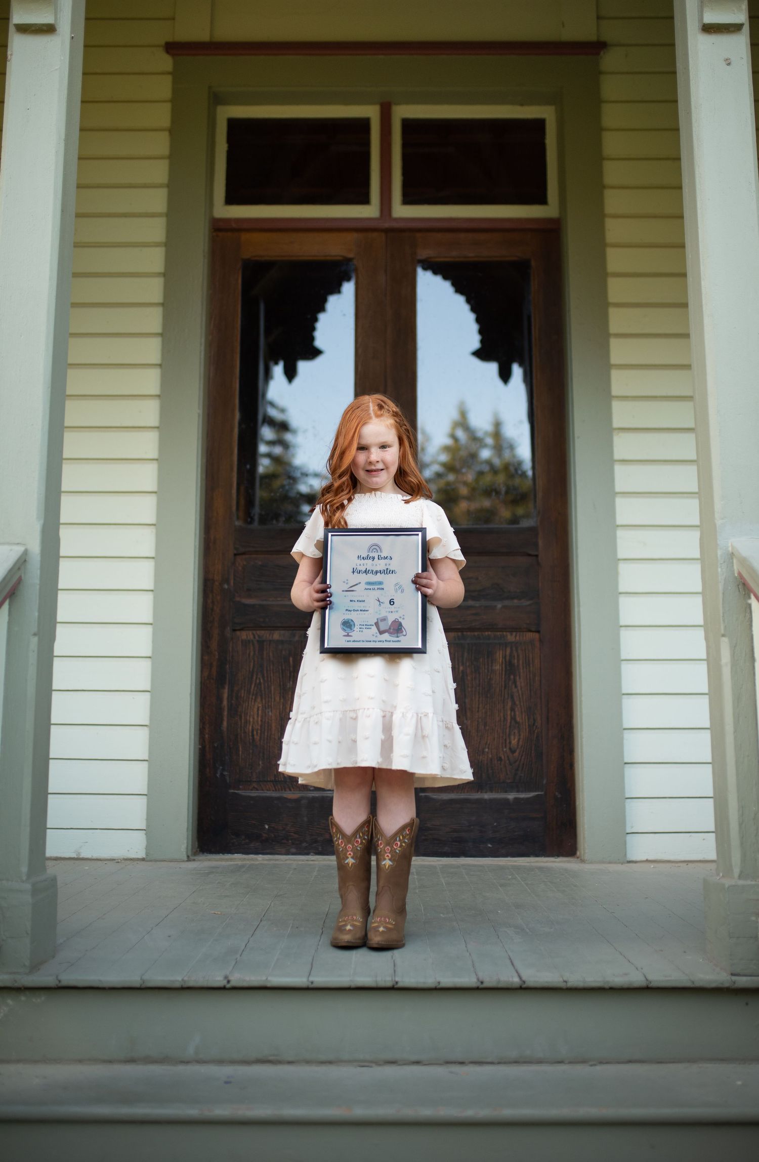 Red-haired girl in white dress and cowboy boots holding a framed certificate on a porch.
