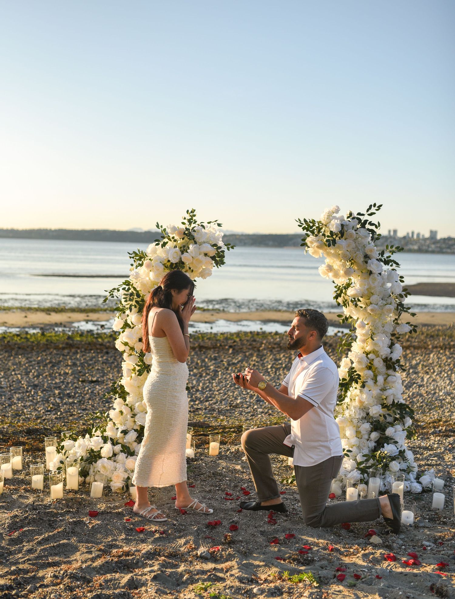 Man proposing on one knee to his partner under a white floral arch on a beach at sunset.