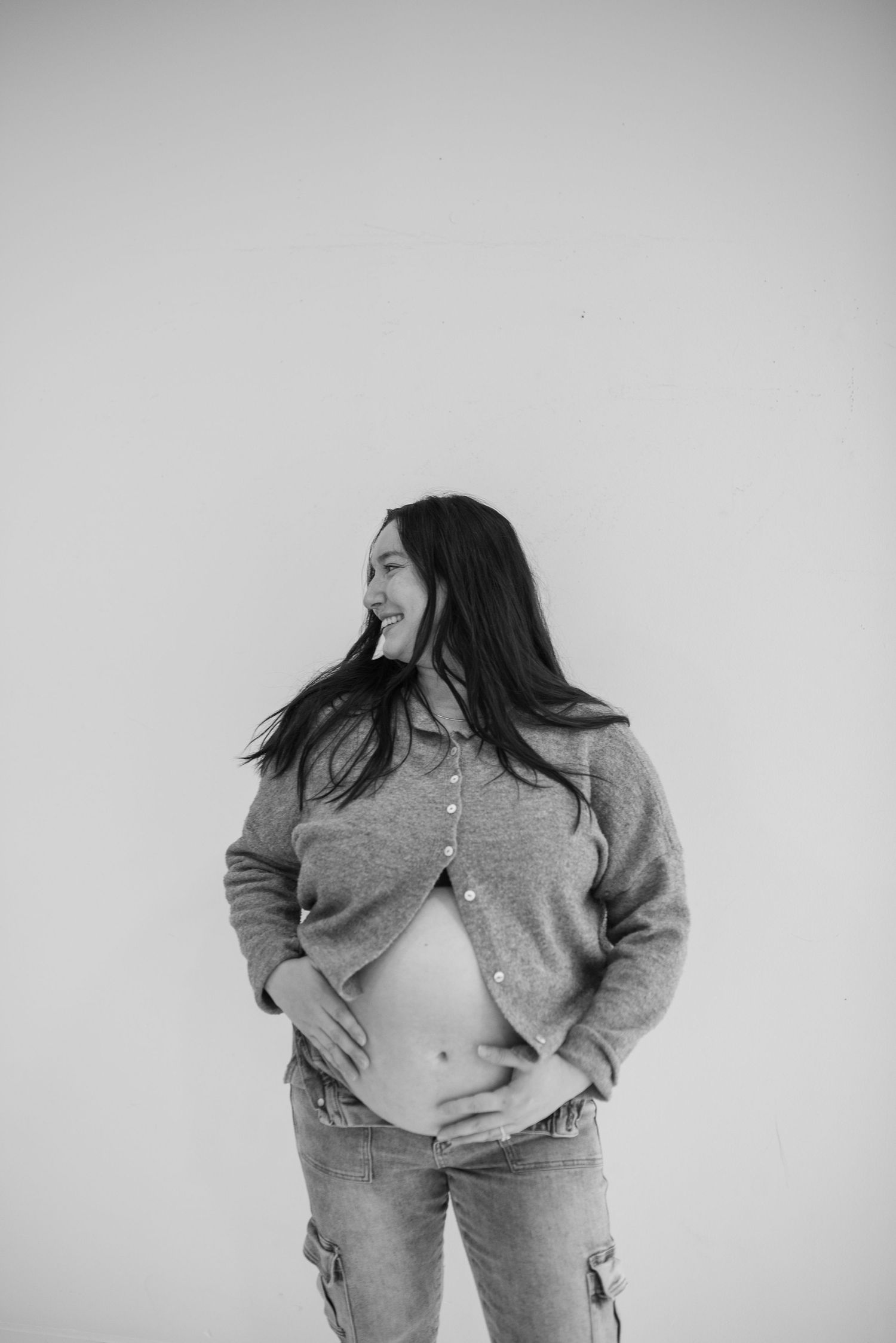 Black and white photo of smiling pregnant woman in open gray cardigan holding her bare baby bump.