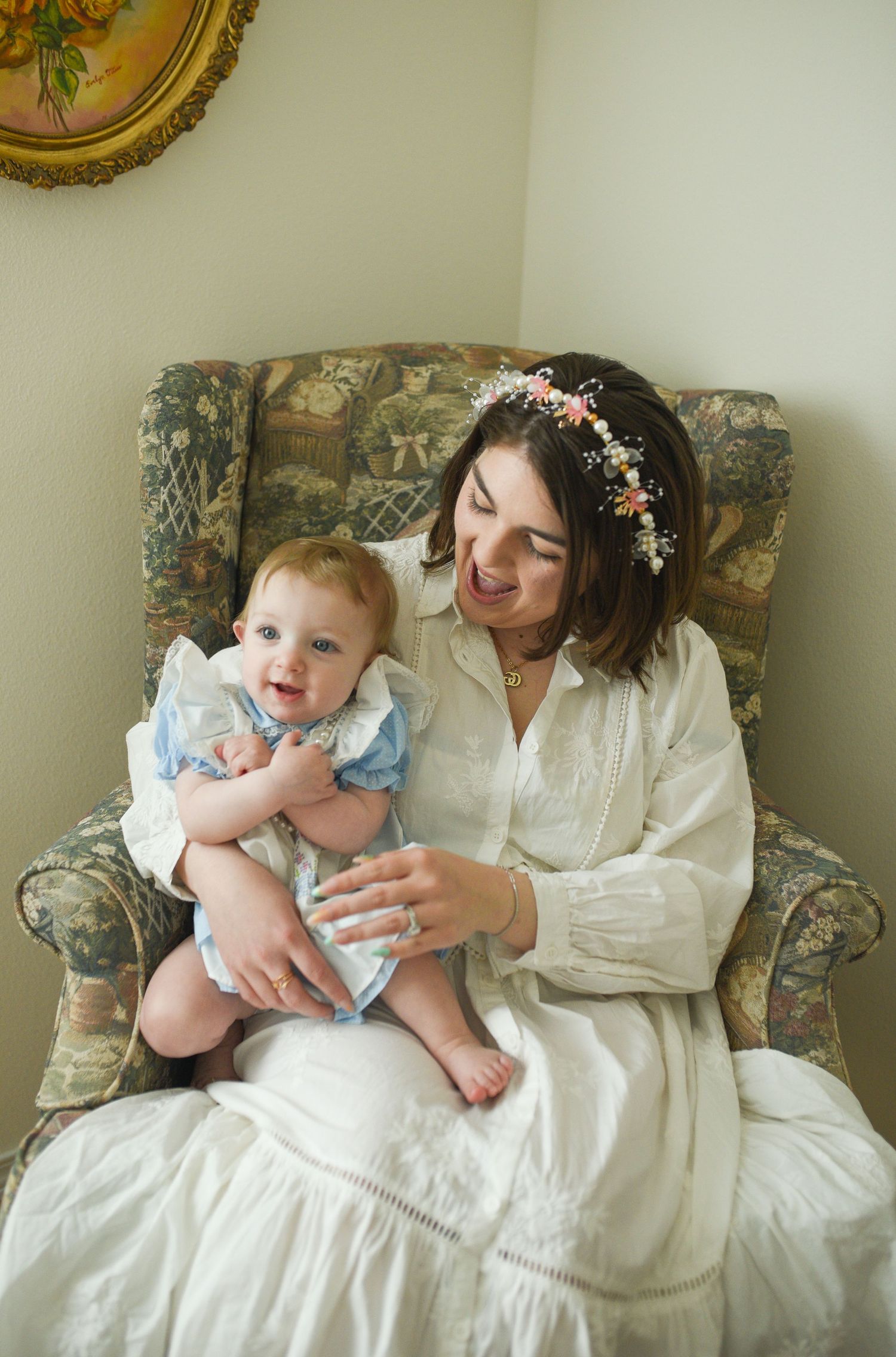 A smiling mother wearing a floral crown sits in a floral armchair holding her baby in a blue dress.