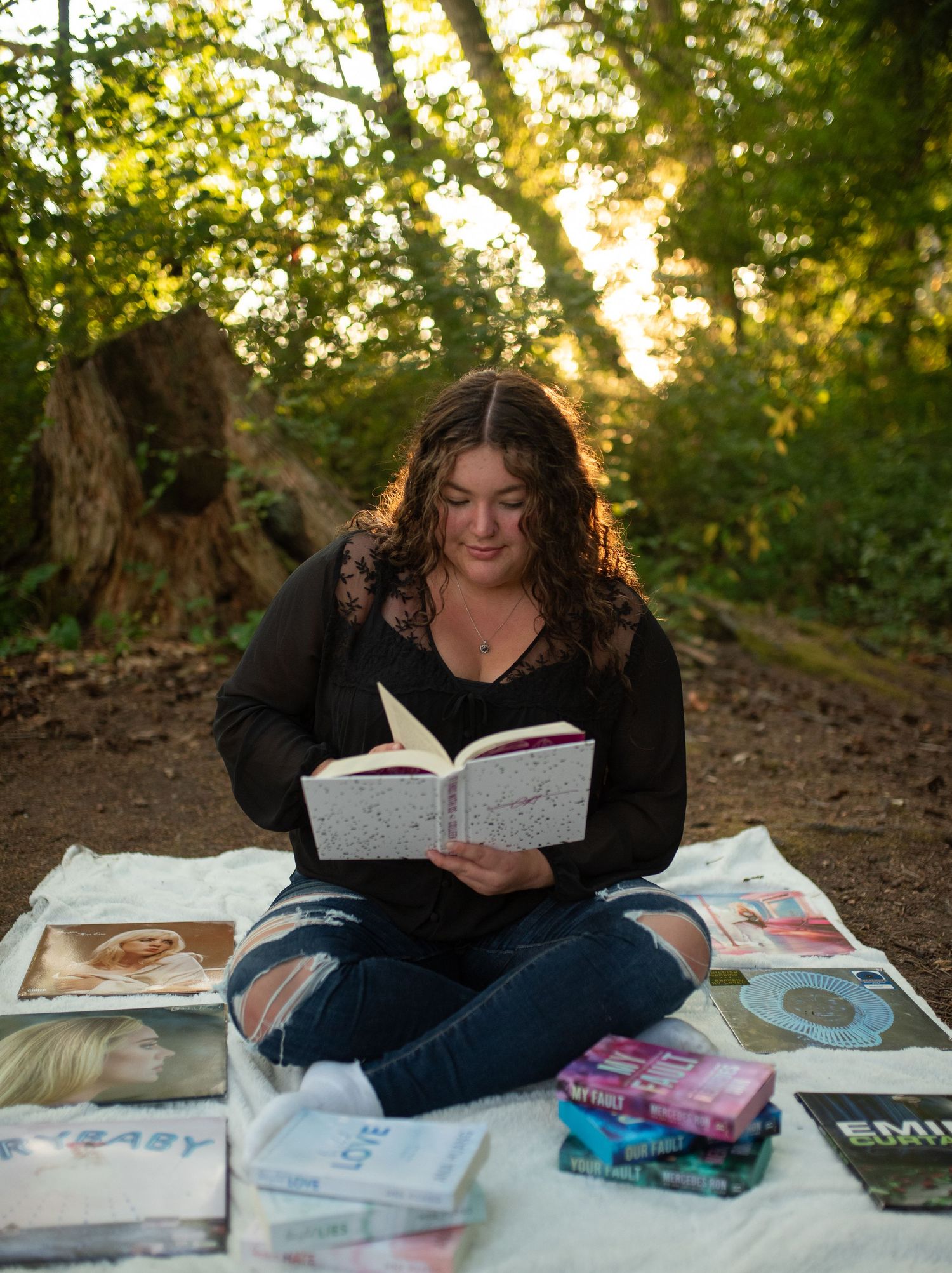 Teen girl reads book sitting on blanket with vinyl records in forest.